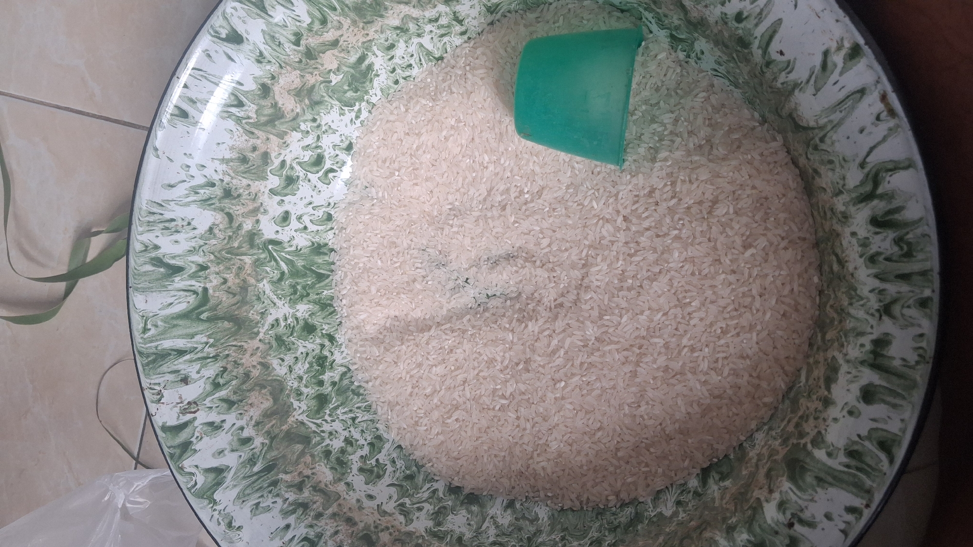 A high-angle, medium shot shows a large quantity of uncooked white rice in a white and green enameled metal basin. The rice fills the majority of the basin, creating a mound in the center. A small, teal-colored plastic scoop is partially submerged in the rice on the right side of the basin. The basin has a distinctive swirled pattern of dark green on a white background. The background consists of a light-colored tiled floor and a corner of a clear plastic bag, with a piece of green ribbon or plant stem draped over the top of the basin. The image is taken in a domestic setting, likely a kitchen or pantry, in Denpasar, Indonesia.