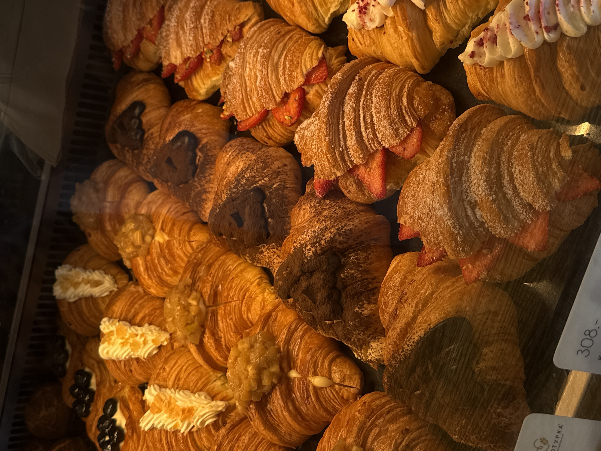 This image shows a display case filled with various croissants in Izhevsk, Russia. The croissants have different toppings: strawberries, cream, chocolate powder, and other sweet toppings. There is a label with the number 308 partially visible on the right side of the frame. The lighting is warm. The croissants are arranged in rows.