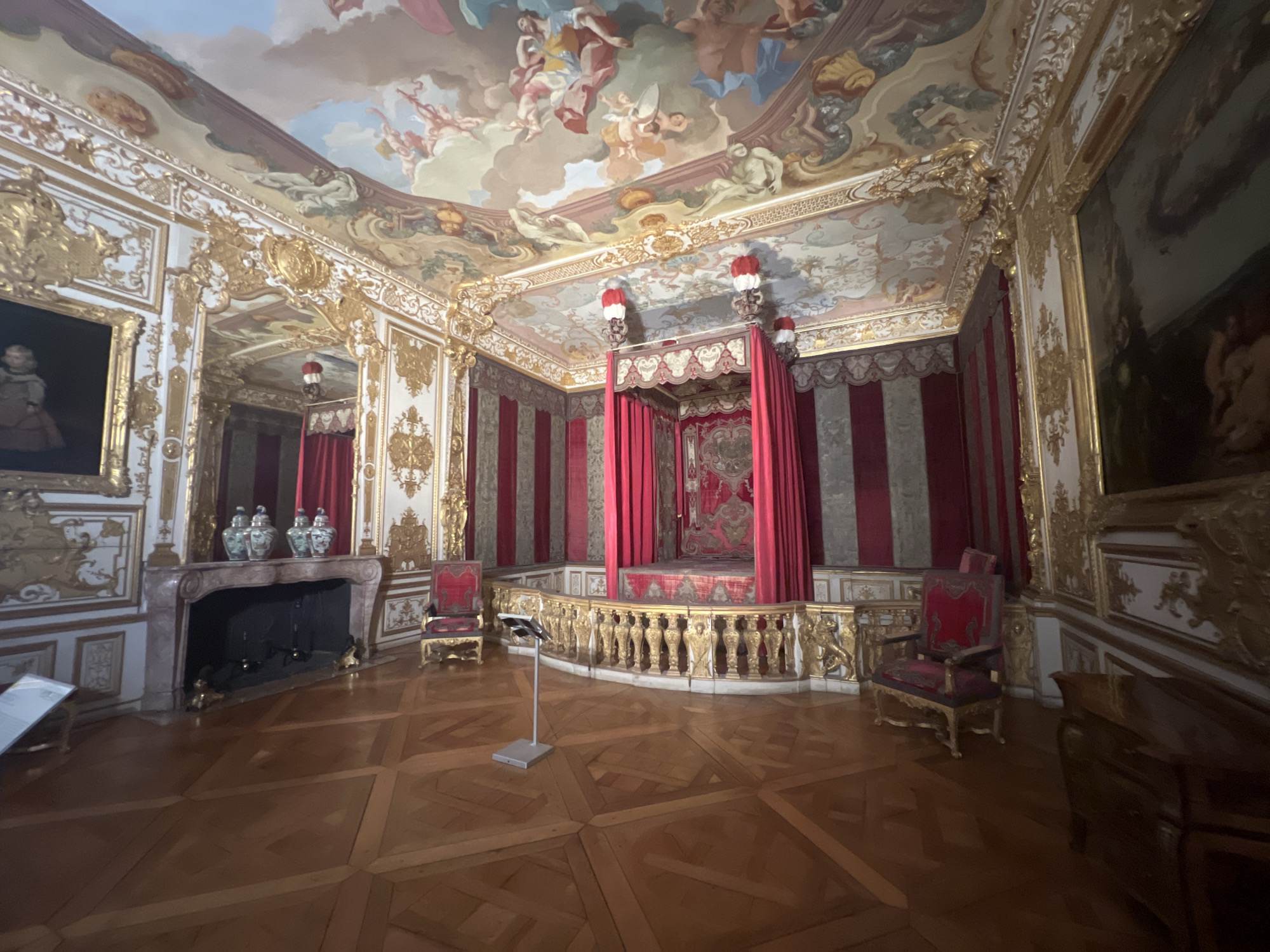 This image depicts the Audience Chamber in the New Palace of Schleissheim, located in Oberschleißheim, Germany. The chamber is opulently decorated with gilded moldings, striped red and grey drapery, and a richly painted ceiling featuring mythological scenes. A grand four-poster bed with red curtains is the focal point of the room, raised on a platform surrounded by a golden balustrade. Two ornate red chairs with gilded accents are positioned on either side of the bed. To the left of the bed, a marble fireplace with a mantelpiece adorned with decorative vases is visible. The floor is made of polished wooden parquet. The lighting suggests daytime. There are no people visible in the image.