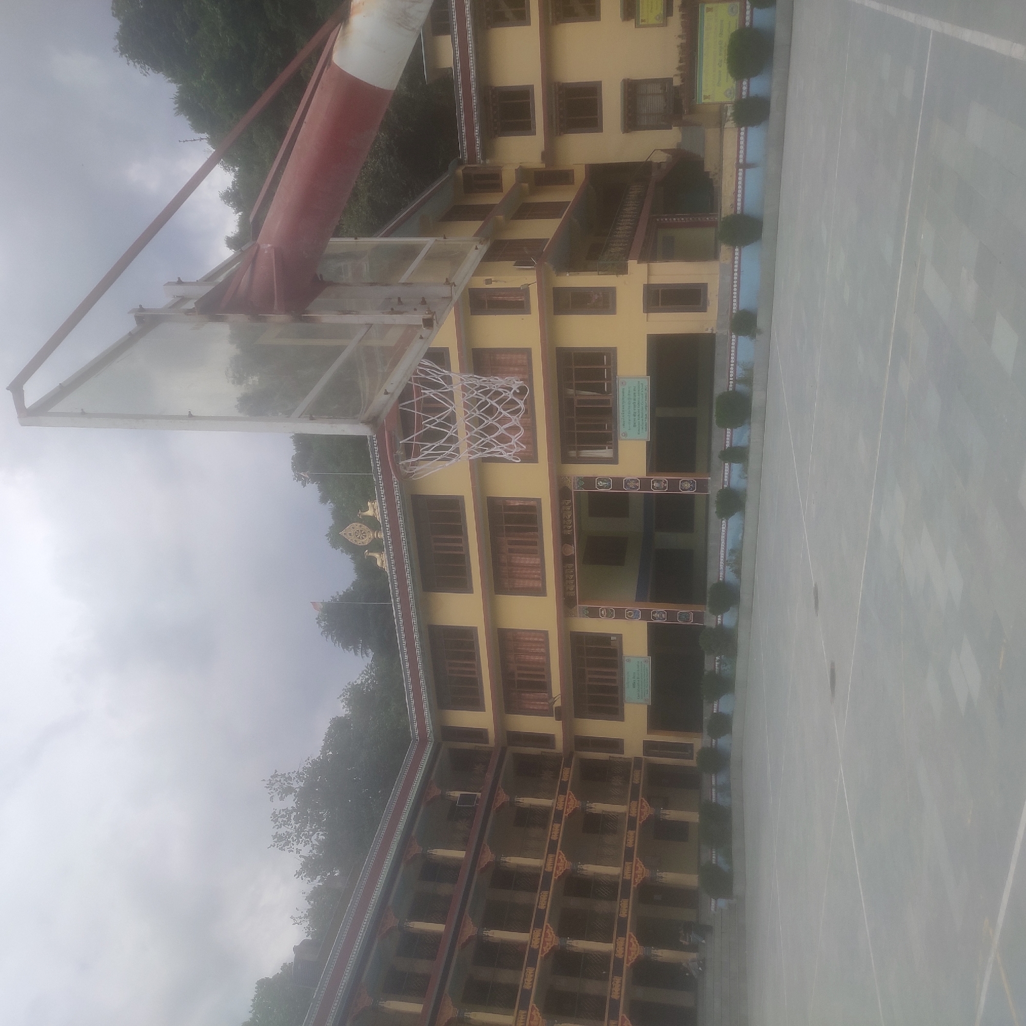 In Batahar, India, a large, yellow, multi-story building with ornate balconies and decorative trim stands as the central subject of the image. The building appears to be a school or cultural institution, possibly a monastery, indicated by the Tibetan-style architecture and the Dharma wheel emblem on its roof. In front of the building, a basketball court is visible, with a basketball hoop and backboard positioned prominently in the foreground, slightly to the right. The court surface is grey with white lines. The sky above is overcast and hazy, suggesting a cloudy or foggy day. Lush green trees and foliage surround the building, with a hillside visible in the background. A few individuals are present, appearing as small figures on the building's steps and the court, but their details are not discernible. The overall scene depicts an educational or cultural complex in a natural, possibly mountainous, setting.
