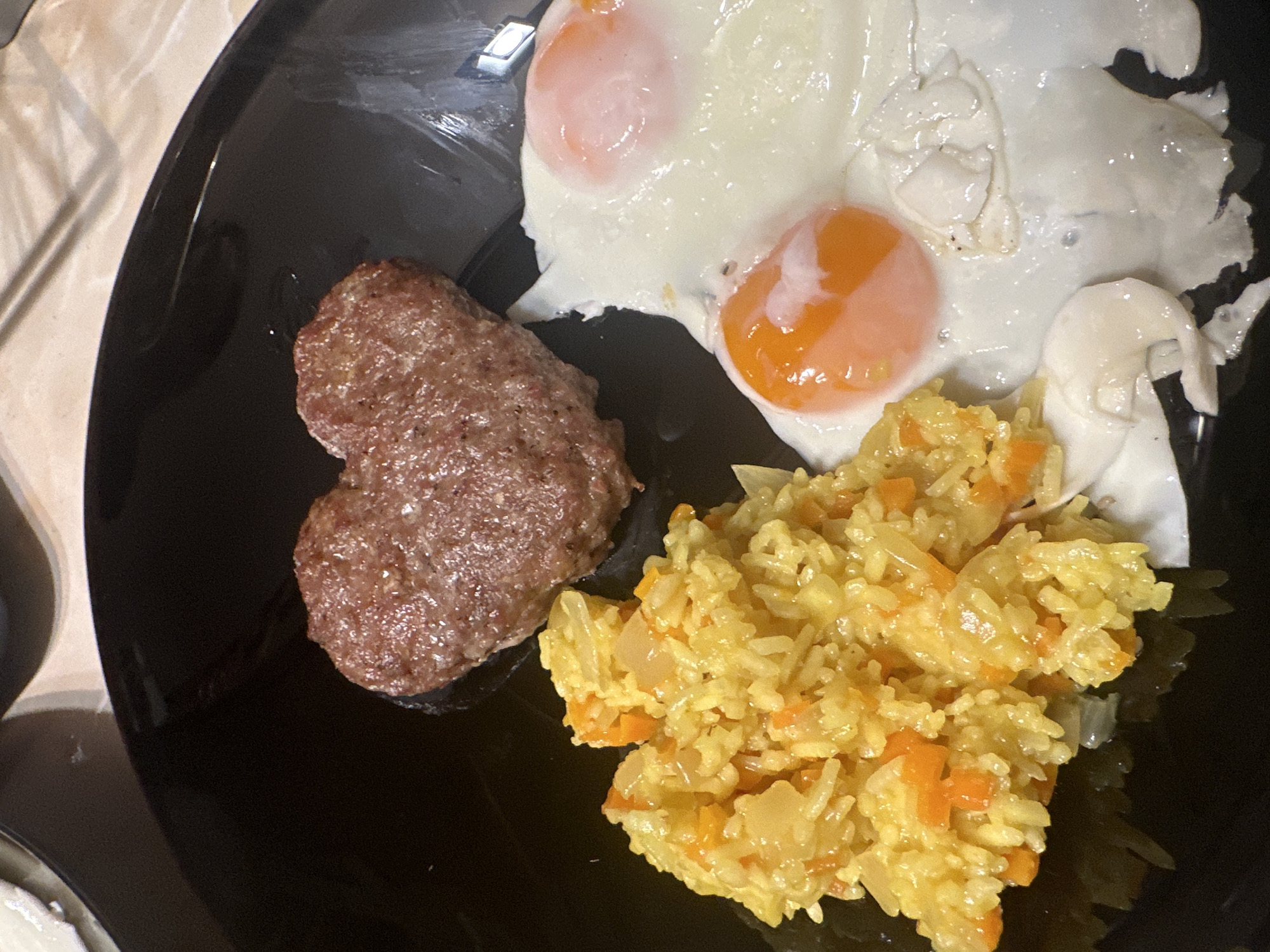 A plate of food is presented, consisting of a piece of cooked meat shaped like a heart, two fried eggs, and a portion of yellow rice with chopped vegetables. The plate is black and glossy, and is likely situated indoors. The location cannot be determined from the image.