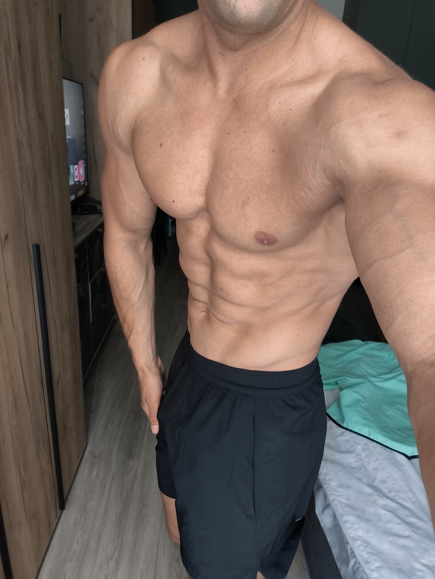 The image shows a close-up of a muscular man's torso and upper legs. He is wearing black athletic shorts. His skin is tanned and he has well-defined abdominal muscles and chest muscles. The background includes a portion of a wooden wardrobe, a television screen, and a bed with light blue and grey linens. The flooring appears to be laminate wood. There are no visible indicators to suggest the location is in Moscow, Russia.