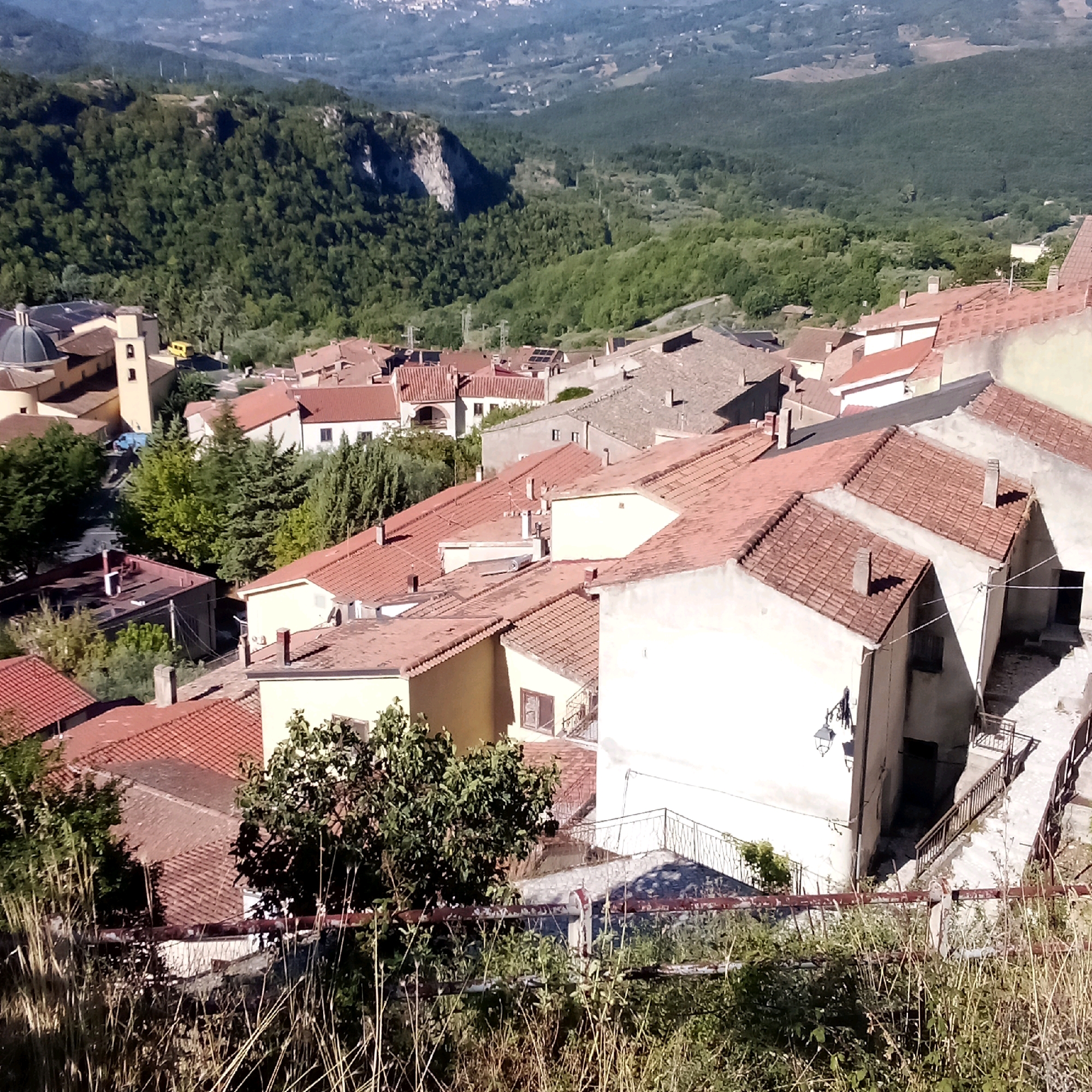 The image presents an elevated perspective of the town of Santomenna, Italy, showcasing its characteristic terracotta-tiled roofs and surrounding verdant landscape. In the foreground, a cluster of buildings with red-tiled roofs dominate the view. These structures, varying in height and architectural style, are densely packed together, suggesting the narrow streets typical of an Italian hill town. Some buildings exhibit pale yellow or white facades, while others are partially obscured by lush green trees and dry grasses in the immediate foreground, which are likely part of the natural environment on the town's edge.
Further back, a prominent church building stands out with its distinctive dome and bell tower, painted in a light yellow hue. This religious structure is a focal point, hinting at the historical and cultural significance of the town. The buildings extend towards the background, where the terrain begins to rise into forested hills. The hills are covered in dense green trees, creating a stark contrast with the built environment. The background reveals a vast, undulating landscape with more hills and valleys, suggesting a rural or mountainous region. A faint haze in the distance indicates the presence of more distant features or atmospheric conditions.
The overall scene is one of a picturesque Italian town nestled within a natural environment, likely captured on a sunny day given the clear shadows cast by the buildings and vegetation. There are no visible people or specific actions taking place in the image, rendering it a still-life depiction of the town and its surroundings. The composition emphasizes the architectural density of Santomenna against the backdrop of its natural landscape.
