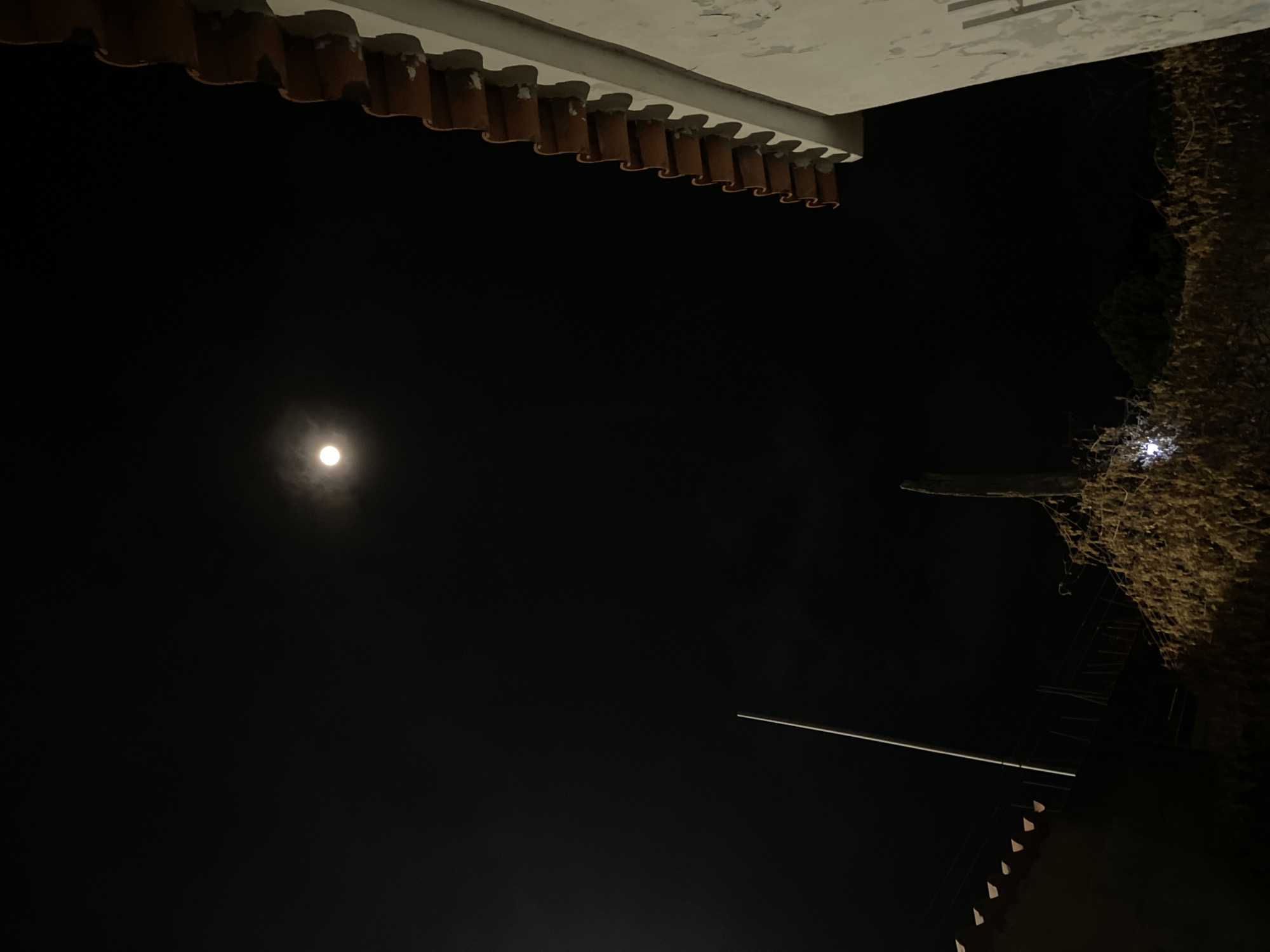 The image shows a night scene in Juan Griego, Venezuela. A full moon is visible in the upper left portion of the frame. The edges of two building structures frame the top portion of the image: a white rooftop structure with reddish-brown corrugated tiles on the left, and a tall exterior wall, covered in vines, on the right. A portion of the wall contains stairs. A thin white electrical conduit wire extends diagonally across the middle-right portion of the dark sky.