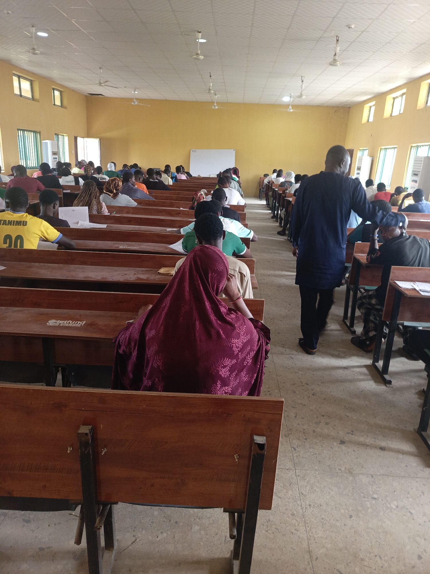 This media file captures an indoor scene within a large hall or classroom in Akwanga, Nigeria, likely during an examination or lecture. Numerous individuals, presumed to be students, are seated at long rows of interconnected wooden desks and benches, facing towards the front of the room.
The hall features light yellow walls and a light-colored ceiling equipped with several ceiling fans. Natural light illuminates the space through multiple windows positioned along the side walls. In the immediate foreground, the back of a person dressed in a dark red patterned garment is visible. To their left, another individual wears a yellow T-shirt with "TANIMU" and "00" printed on the back, seated at a desk with papers. Most of the other seated individuals are seen from behind or from a distance, appearing focused on their desks or the front of the room.
A person in a dark blue tunic, likely an invigilator or instructor, is seen walking down one of the aisles, observing the proceedings. Papers are present on several desks. Notable text includes "TANIMU" and "00" on a yellow shirt, and partial text "Steffund" and "Online Information" visible on the back of one of the wooden desks. A whiteboard or screen is positioned at the far end of the hall. The scene depicts a quiet, formal academic setting during daylight hours.