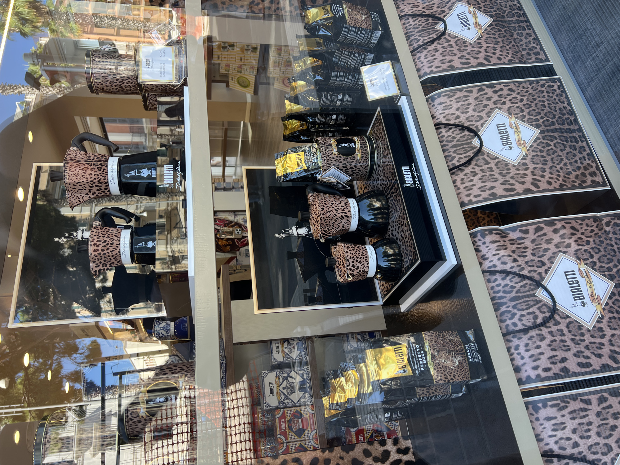 This image shows a display window of a store in Bari, Italy, featuring Bialetti coffee products with a leopard print design. The display includes several Bialetti Moka Express coffee pots, coffee bags, and shopping bags, all adorned with a leopard print pattern. Some of the coffee pots are black with leopard print tops, and others appear to be entirely made of leopard print material. Several bags of Bialetti coffee are stacked in the window, with gold accents on their packaging. The shopping bags at the bottom of the display are also made of leopard print material and feature the Bialetti logo. In the background, reflections of the street and buildings in Bari can be seen, including palm trees and architectural details characteristic of the city. The overall impression is one of a stylish retail display promoting a limited-edition collaboration.