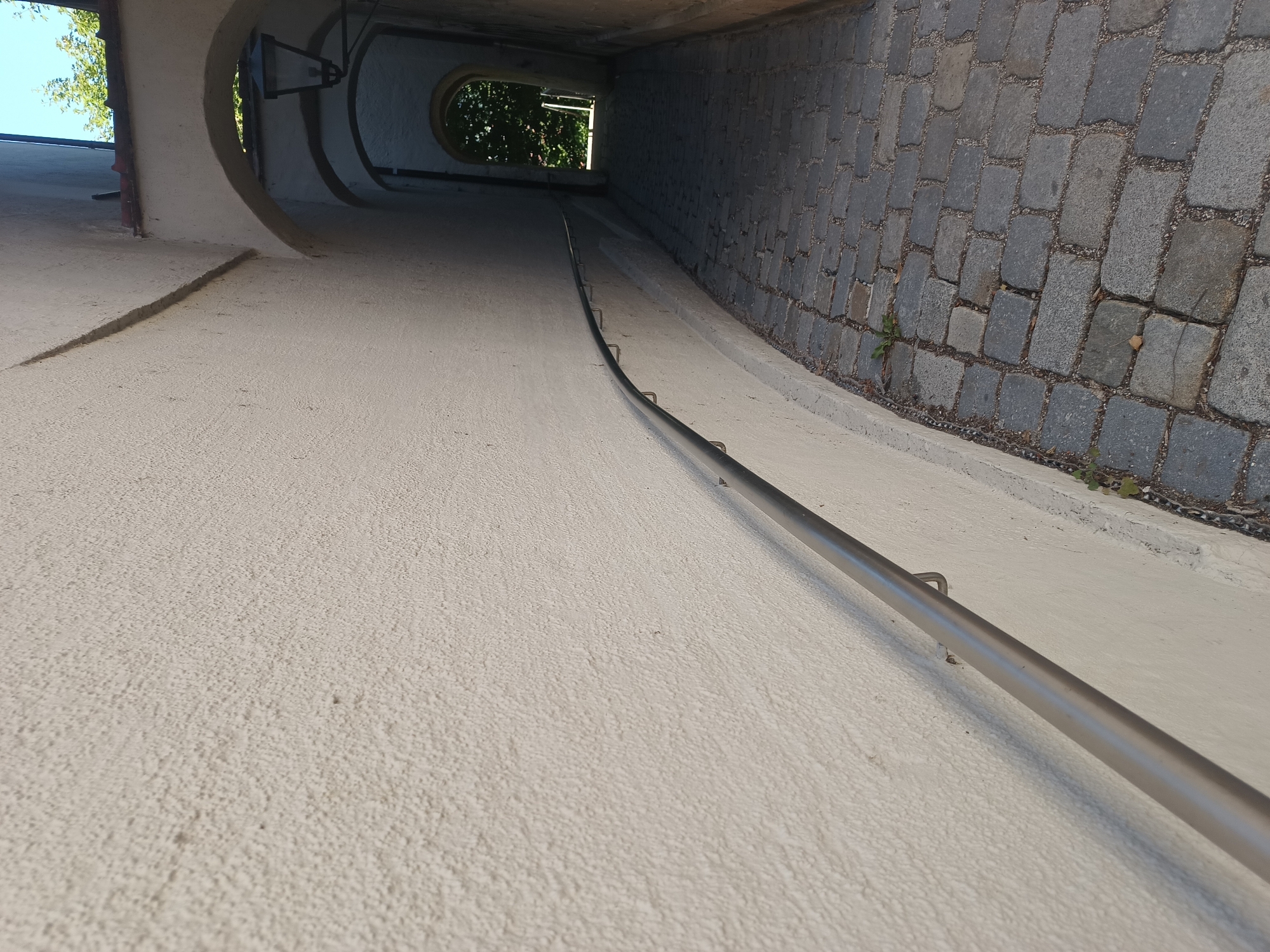 The image shows a cobblestone alleyway in the city of Neuötting, Germany. The alley is lined with a textured, cream-colored wall on the left, which has a metal handrail attached. The right side of the alley is a series of arched passageways. The ground is paved with grey cobblestones. Trees and foliage are visible through the furthest archway, suggesting an outdoor area beyond. The lighting suggests it is daytime, with shadows cast by the arches. There are no people or animals visible in the image.