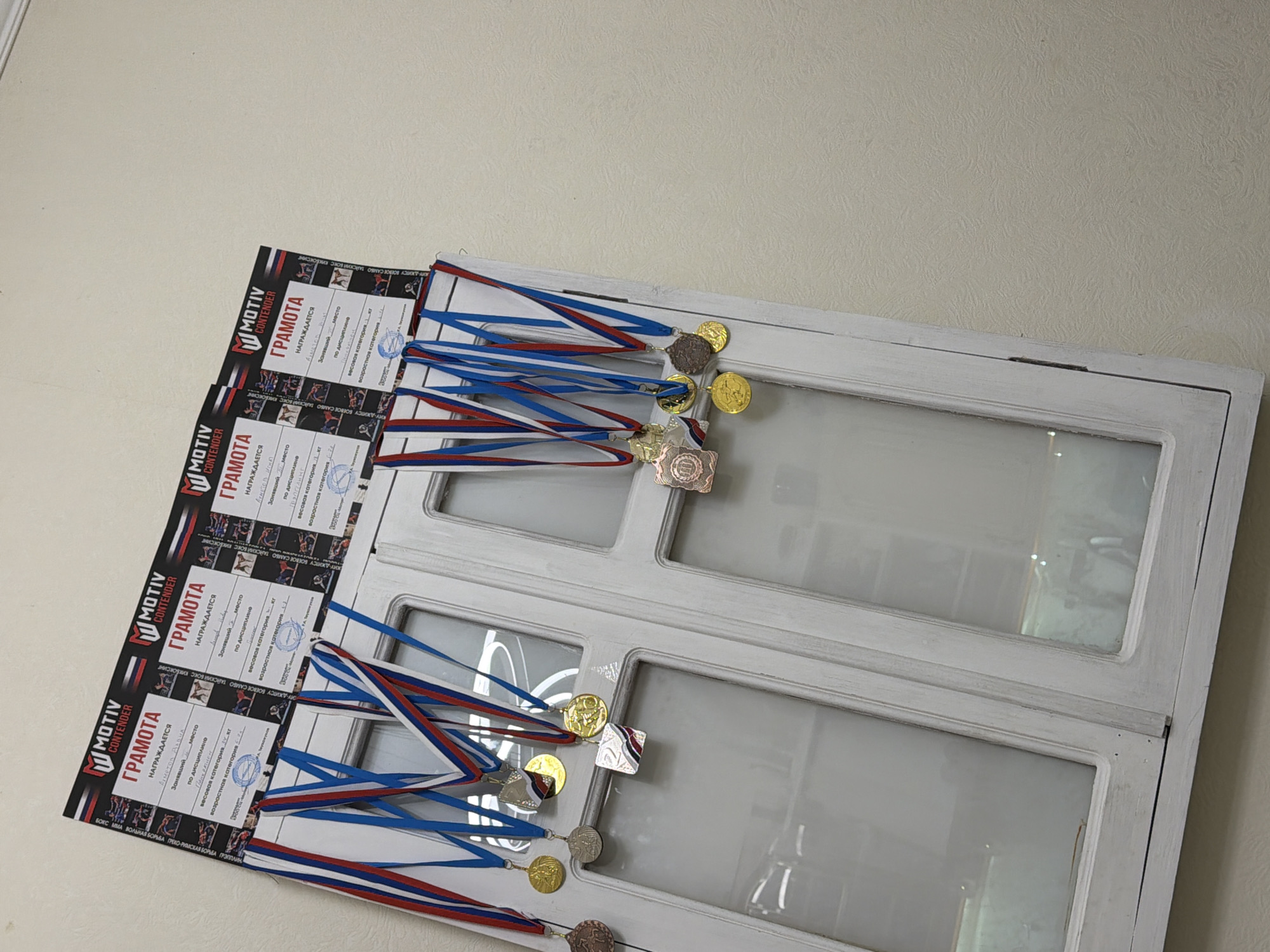 The image captures four award certificates, each prominently labeled "MOTIV CONTENDER ГРАМОТА" (Diploma), laid out horizontally against a light-colored, textured wall. Each certificate is paired with several medals, featuring red, white, and blue ribbons, colors typically associated with the Russian flag. The medals display gold, silver, and bronze finishes, signifying various levels of achievement.
The text on the certificates, in Russian, includes "НАГРАЖДАЕТСЯ" (Awarded to), a space for "Занявший ___ место" (Took ___ place), "по дисциплине" (in discipline), "весовая категория ___ кг" (weight category ___ kg), and "возрастная категория" (age category). One partially visible name is "Ахметов ДаQUA". The disciplines listed on the certificates span a range of combat sports, including "БОКС ММА ВОЛЬНАЯ БОРЬБА ГРЕКО-РИМСКАЯ БОРЬБА ГРЭППЛИНГ" (Boxing, MMA, Freestyle Wrestling, Greco-Roman Wrestling, Grappling), and "ЖИТСУ БОЕВОЕ САМБО ТАЙСКИЙ БОКС КИКБОКСИНГ" (Jujutsu, Combat Sambo, Muay Thai, Kickboxing).
These awards are displayed on what appears to be an old, white-painted wooden frame, possibly a door or window, with glass inserts, resting against the wall. The scene suggests a collection of accolades from a martial arts or combat sports competition, likely in Sochi, Russia, given the provided context. The lighting indicates an indoor setting.