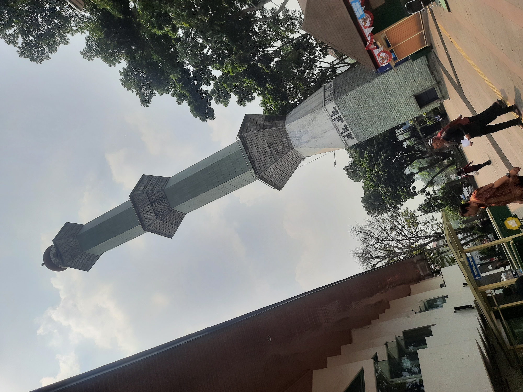 A tall, green, octagonal minaret with a dome at its peak dominates the upper right quadrant of the image, leaning dramatically to the left. The minaret is constructed in sections, with decorative bands of geometric patterns between each level. The lower portion of the minaret is wider and appears to be made of stone or brick. To the left of the minaret, a modern, white building with a dark brown, sloped roof is visible. The building has multiple rectangular windows. In the foreground, a paved area with yellow lines marking a path is visible. Several people are walking in this area. On the far right, a small building with a brown roof and decorative flags hangs down. The sky is overcast with scattered clouds. The scene appears to be outdoors in a city environment, potentially a religious or cultural complex. The specific location in Bandung City, Indonesia, is the Masjid Raya Bandung, specifically its prominent minaret.