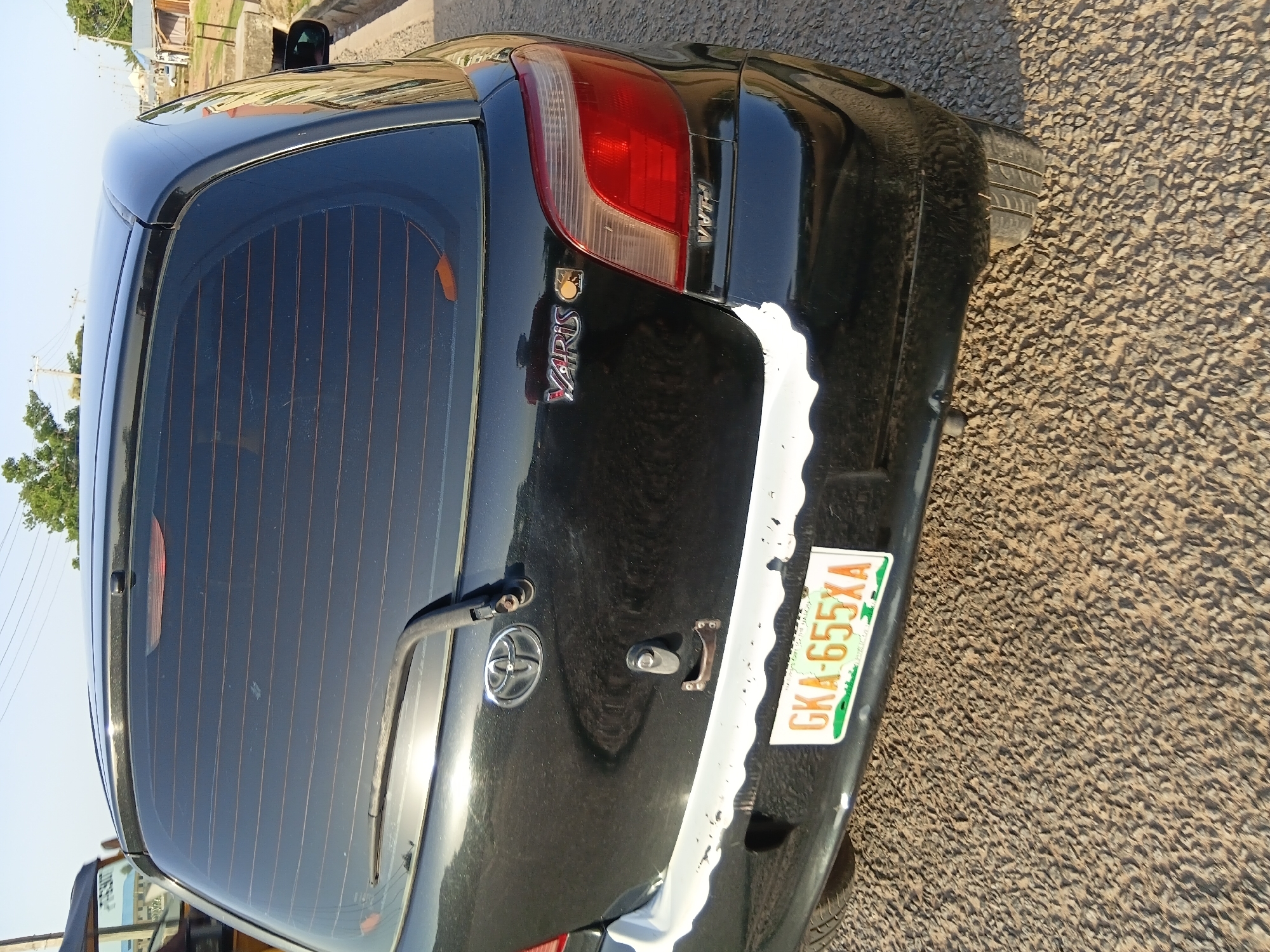 A black Toyota Yaris is parked on a gravel road in Jalingo, Nigeria. The rear of the car is visible, showing the taillights, a license plate that reads "GKA-655XA", and a Toyota emblem. The bumper appears to be painted white and shows signs of wear. The background includes some vegetation and buildings.
