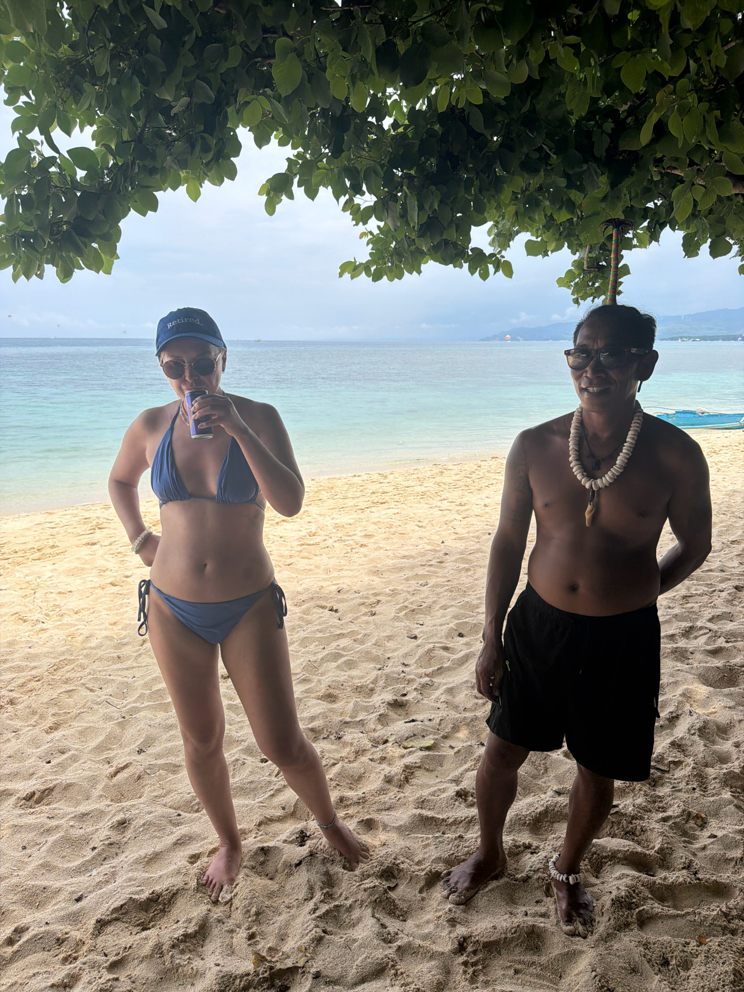 Two people are standing on a sandy beach in Malay, Philippines, under the shade of a tree. On the left, a woman wearing a blue bikini and a blue "Retired" baseball cap is holding a can to her mouth. She has sunglasses on and a white bracelet on her left wrist. On the right, a man with sunglasses and a shell necklace is shirtless and wearing black shorts. He has a small decorative item stuck in his hair. The ocean is visible behind them, with calm turquoise water and a distant shoreline. The sand is light brown and textured with small ripples.