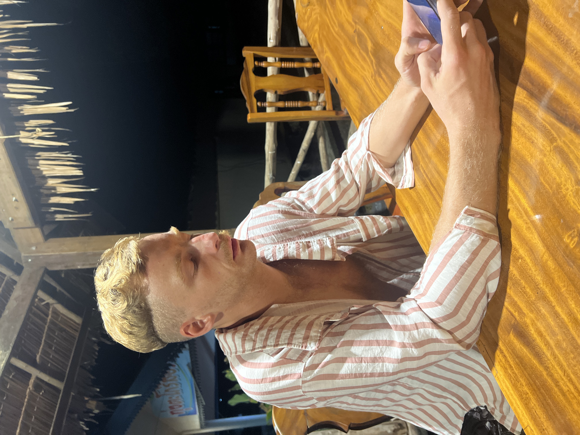 An overhead shot shows a man with blonde, wavy hair and a mustache sitting at a wooden table. He is wearing a pink and white striped button-down shirt, unbuttoned at the top, revealing his chest. The man is looking down at a smartphone in his hands. The background is dark, but a thatched roof and a wooden chair are visible. The image appears to have been taken at night at a restaurant in Thung Tako, Thailand, possibly on a beach due to the thatched roof structure.