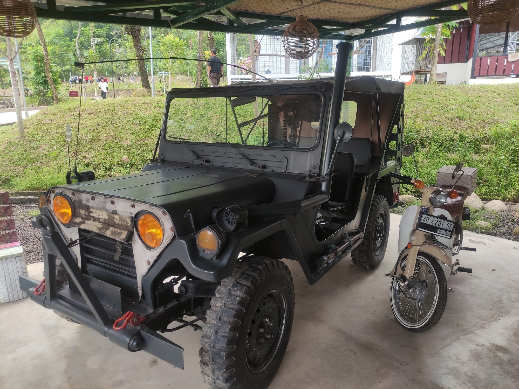 The image shows a gray military jeep and a beige motorcycle parked under a covered area, likely a garage or carport. The jeep has distinctive features such as large, knobby tires, a snorkel exhaust pipe, and a wooden-looking front panel with prominent headlights. The motorcycle, with a license plate reading "AB 4201 GS," is positioned to the right of the jeep. In the background, there's a grassy hill, trees, and a building with a light-colored facade. There are people visible in the distance on the grassy hill. The location is identified as Babakan Madang, Indonesia. A woven lampshade hangs from the ceiling of the covered area. The setting appears to be a rural or suburban environment with greenery.