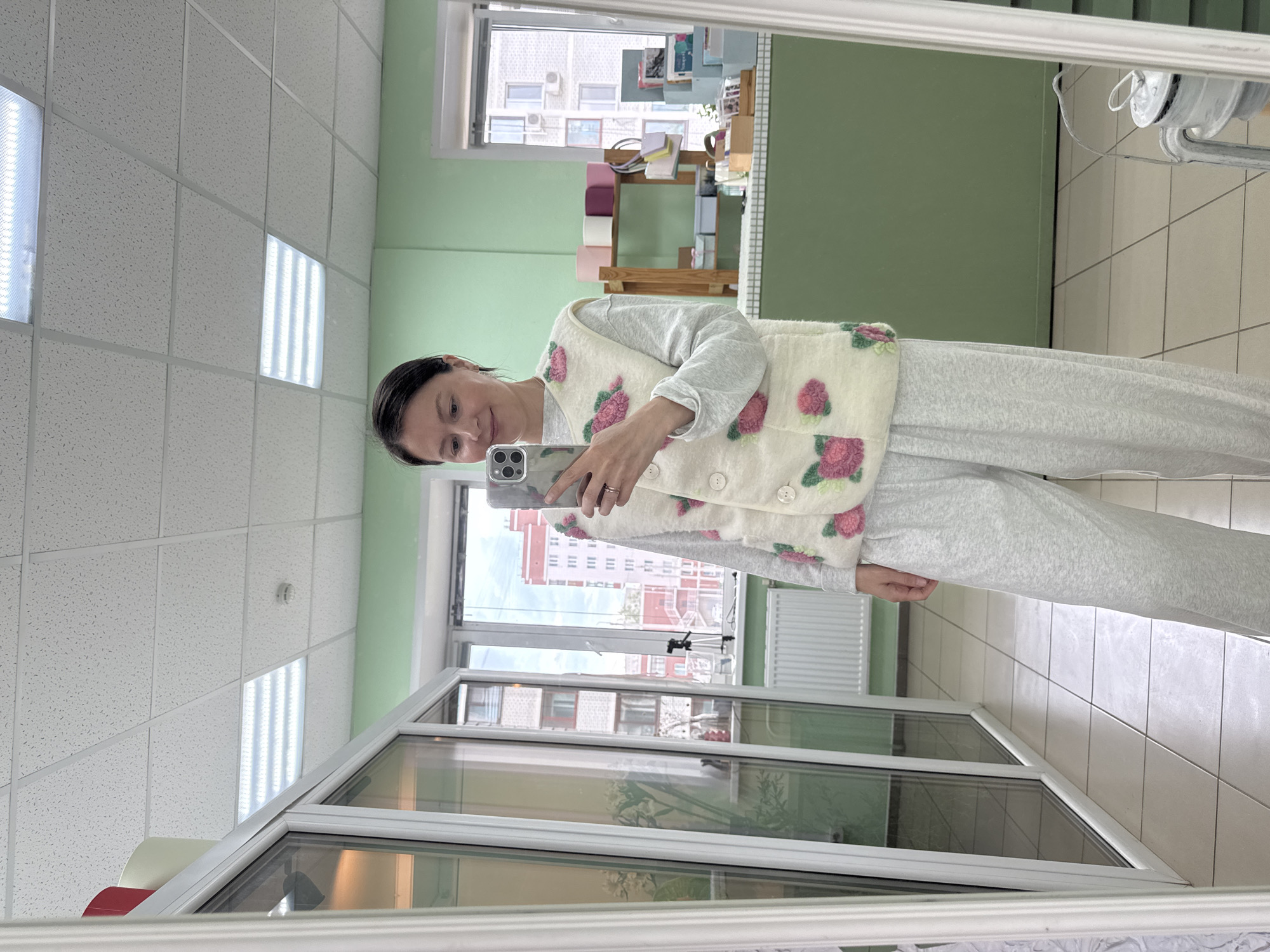 A mirror selfie captures a woman standing in an indoor setting, likely in Izhevsk, Russia, as she looks down at her smartphone. She has dark hair pulled back and displays a soft, content smile.
The woman is wearing a light-colored, possibly cream or off-white, sleeveless vest adorned with distinct pink and green floral appliqués or embroidery. Beneath the vest, she wears a light grey long-sleeved top and matching loose-fitting grey pants, suggesting comfortable attire. A ring is visible on her left hand as she holds a smartphone, which appears to be an iPhone, encased in a clear, patterned cover.
The background, a reflection in the mirror she is using, reveals the characteristics of the room and the exterior. The room features light green walls and a suspended ceiling composed of white rectangular acoustic tiles with integrated fluorescent light panels. A large window with white frames dominates a section of the wall, offering a view of an urban landscape under an overcast sky. Visible through the window are several multi-story buildings, including one with a reddish-orange facade and another grey one, some showing exterior air conditioning units.
Within the reflected interior space, behind the woman, a wooden shelving unit is visible, holding various items such as colorful rolls of fabric or paper, books, and other decorative objects, hinting at a creative studio, craft shop, or office environment. A white radiator is also seen beneath the window. The floor is tiled with light-colored, possibly cream, square tiles. The overall lighting is bright, a mix of natural light from the window and artificial illumination from the ceiling fixtures. No legible text is visible in the image.