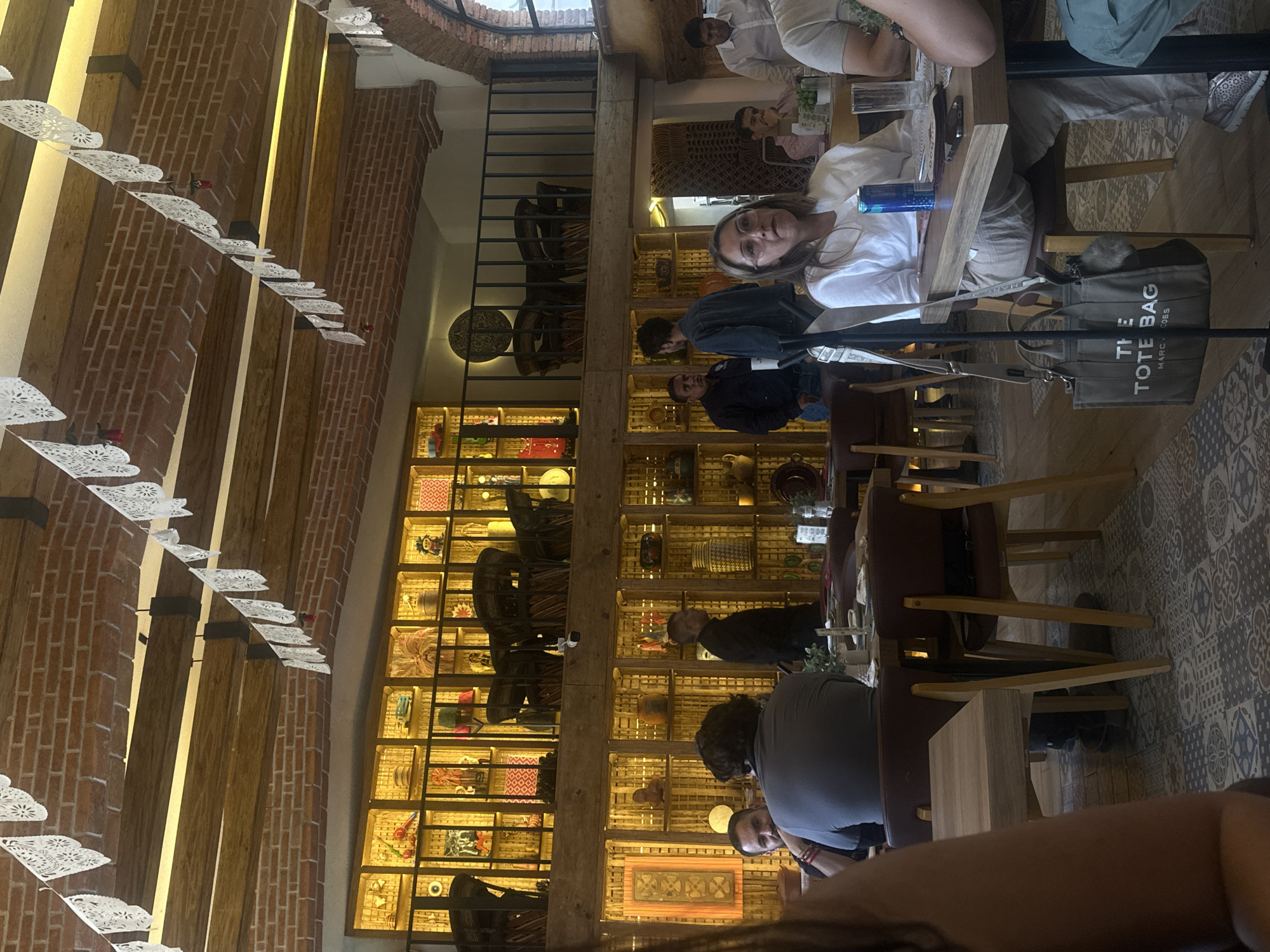 The image captures an interior scene in San Martín de las Pirámides, Mexico. The location appears to be a restaurant or eatery.
People are seated at tables, engaged in conversations and dining activities. There are men and women of varying ages present.
The setting is decorated with traditional Mexican elements, including paper decorations suspended from the ceiling. A prominent wall features a display of shelves filled with various artisanal objects.
A "The Tote Bag" branded tote bag is visible near one of the tables. The scene suggests a casual dining environment in a place of cultural significance.