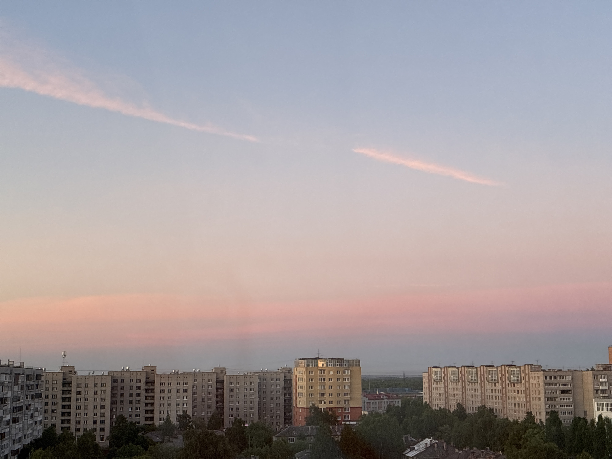 The image is an eye-level, wide-angle shot of a city skyline at dusk. The sky is a gradient of pale blue at the top transitioning to soft pink and peach tones near the horizon, with two thin, pink contrails visible in the upper portion. Below the sky, a row of multi-story apartment buildings spans the width of the frame. The buildings are of varying heights and architectural styles, mostly appearing to be Soviet-era panel buildings. They are grey and beige, with numerous windows. Dense green trees are visible in the foreground, partially obscuring the lower floors of some buildings. The overall impression is a somewhat subdued urban landscape illuminated by the soft light of sunset.