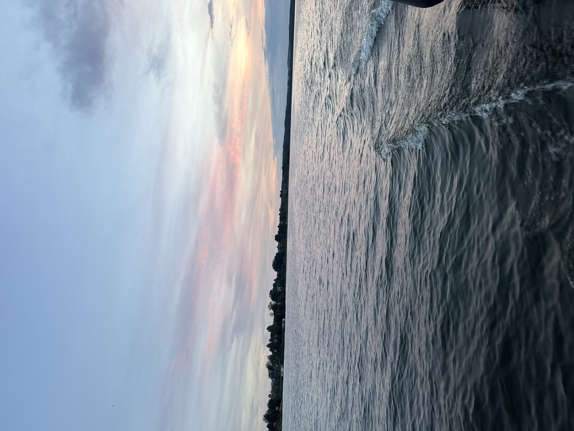The image depicts a serene view of a body of water, likely a lake or river, with a distant shoreline dotted with trees and small structures. The sky above is a gradient of soft blues and pinks, suggesting either dawn or dusk. The water is calm with gentle ripples, and a visible wake emanates from the right side of the frame, indicating movement, possibly from a boat. The overall atmosphere is peaceful and natural. The scene is consistent with the Votkinsk Reservoir, a large artificial lake on the Kama River located near the city of Votkinsk in Russia, known for its picturesque natural surroundings. There are no people or animals visible in the image.