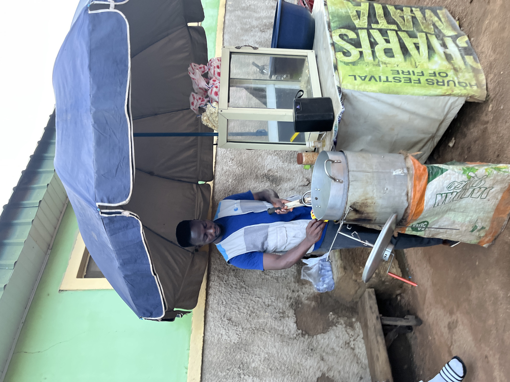 An outdoor scene in Jos, Nigeria, features a man operating a street food stall, possibly grilling or roasting. He is positioned in the center, wearing a blue t-shirt with a light grey, mesh-paneled vest over it, and dark trousers. He appears focused on his task, holding tongs over a large, cylindrical metal pot or drum, which serves as the cooking apparatus. This pot is supported by what looks like a white sack, with additional metal framework holding a dish-like component with a red handle below it.
The stall is covered by a large, dark blue umbrella, providing shade. To the man's right, there is a small, light-colored glass display case, likely for showcasing food items. Next to this display case, a black box (possibly a battery pack or speaker) sits on a makeshift table. A blue basin and several white plastic bags with red markings are also visible in the background, suggesting supplies or packaged goods.
Behind the stall, a banner is draped, displaying prominent text: "SHARIS MATA" and "HOURS FESTIVAL OF FIRE." Another white sack with some visible text, possibly "MAIDAN," is part of the support structure for the cooking setup. The ground is unpaved, appearing as dirt or a rough concrete surface. In the background, a building with a light green wall and a window is partially visible, indicating a settled area. The overall lighting suggests it is daytime.