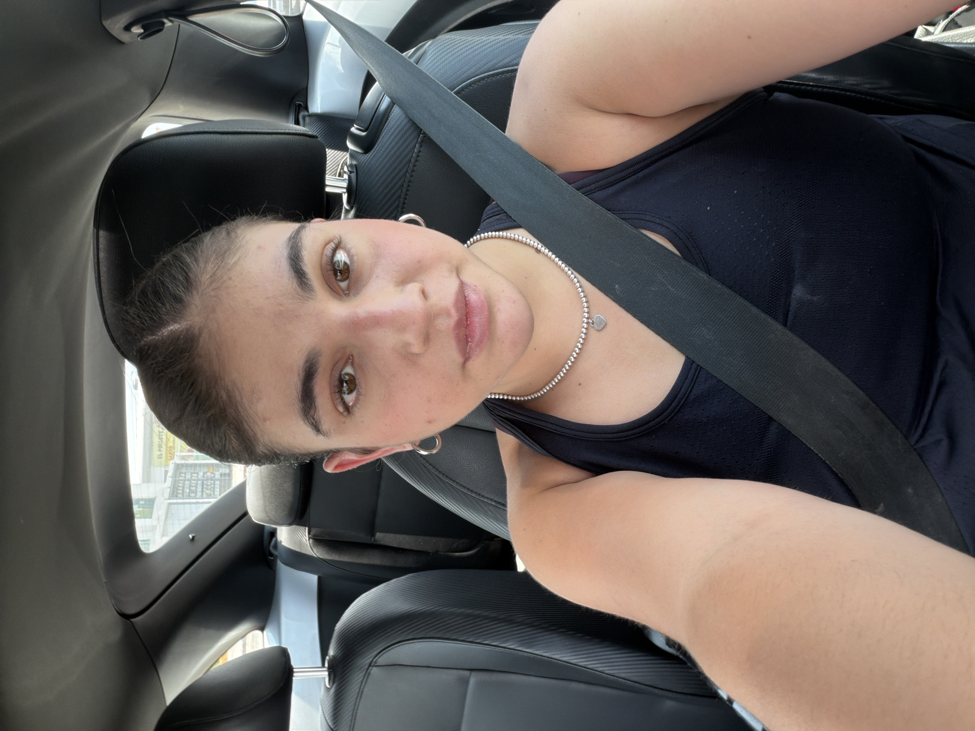 This image is a rotated selfie taken by a young woman inside a car during the daytime. The original image is oriented 90 degrees clockwise.
The main subject is a young woman, likely in her late teens or early twenties, with brown eyes and dark hair pulled back from her face. She has a neutral to slightly smiling expression as she looks directly at the camera. She is wearing a black tank top and a silver beaded necklace with a small, heart-shaped charm. Silver hoop earrings are visible on her ears. A black seatbelt is diagonally secured across her chest and shoulder.
The setting is the interior of a modern vehicle. Visible elements of the car include black leather seats, some with a textured pattern, a black dashboard or door panel, and a dark grey interior roof lining. Bright natural light illuminates the scene, indicating it is daytime. Through the car window, a blurry urban or suburban background can be seen, featuring buildings. In one of the cropped images, faint, blurry text "EL PR" is visible on an outdoor structure. The activity is a casual self-portrait.
The location context provided is San Bernardino Tlaxcalancingo, Mexico.