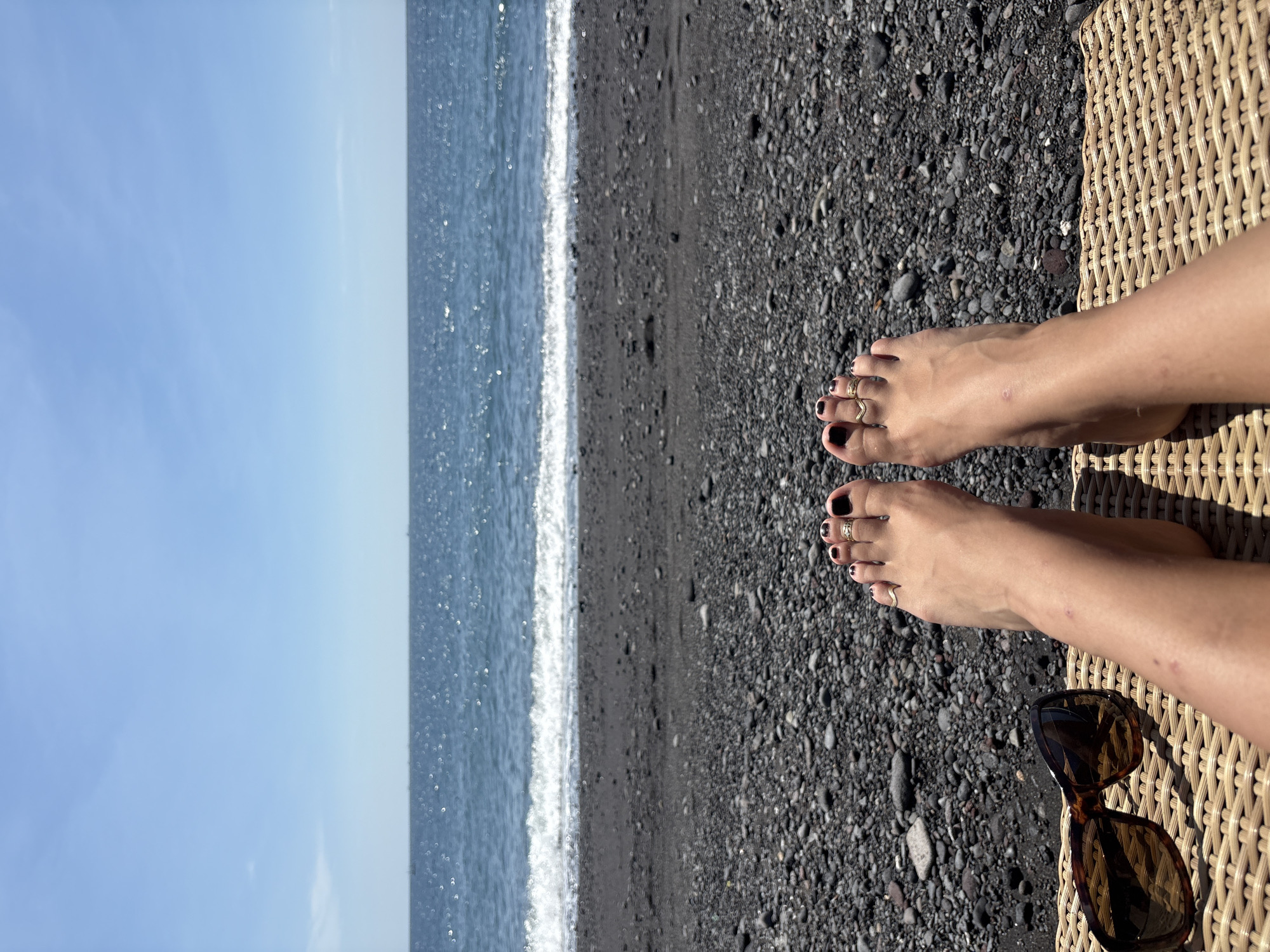 A person's feet are positioned in the foreground, resting on a woven surface, possibly a beach chair. The feet have black nail polish and are adorned with two rings on the right foot. Next to the feet are a pair of brown-tinted sunglasses. The background reveals a black pebble beach leading to a blue ocean under a clear sky. The horizon line separates the water and the sky. The image appears to be taken outdoors in a sunny environment.