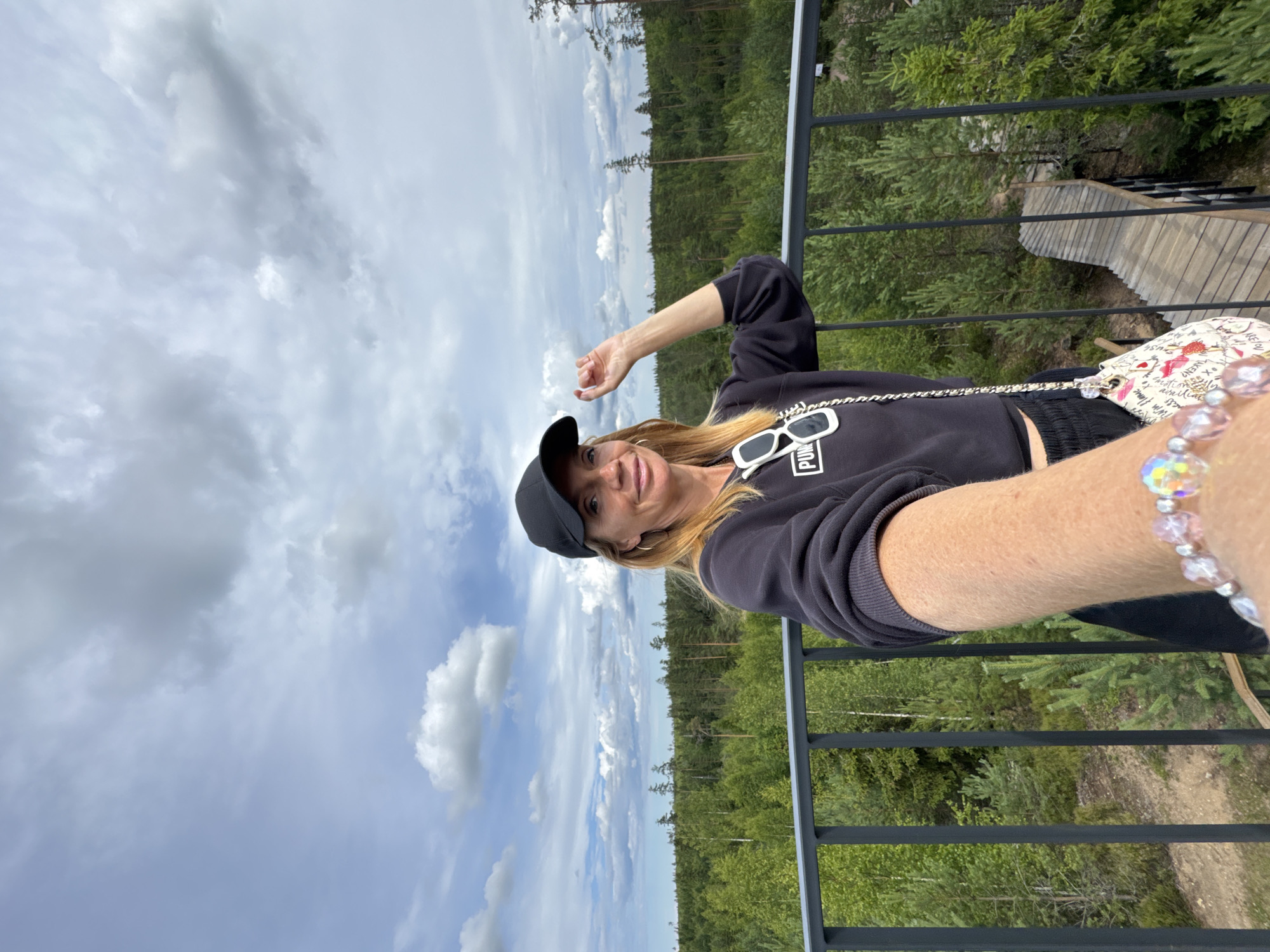 A woman with blonde hair, wearing a black baseball cap, black sweatshirt, and black shorts, stands on an elevated platform overlooking a forest. She is holding her phone out to take a selfie. She has white sunglasses hanging from the neck of her sweatshirt and a patterned purse slung over her shoulder. A beaded bracelet is visible on her left wrist. The background shows a dense green forest with scattered tall trees under a cloudy sky. To the right of the woman, a wooden walkway with railings descends into the forest. The location is the Peski nature reserve in Russia.