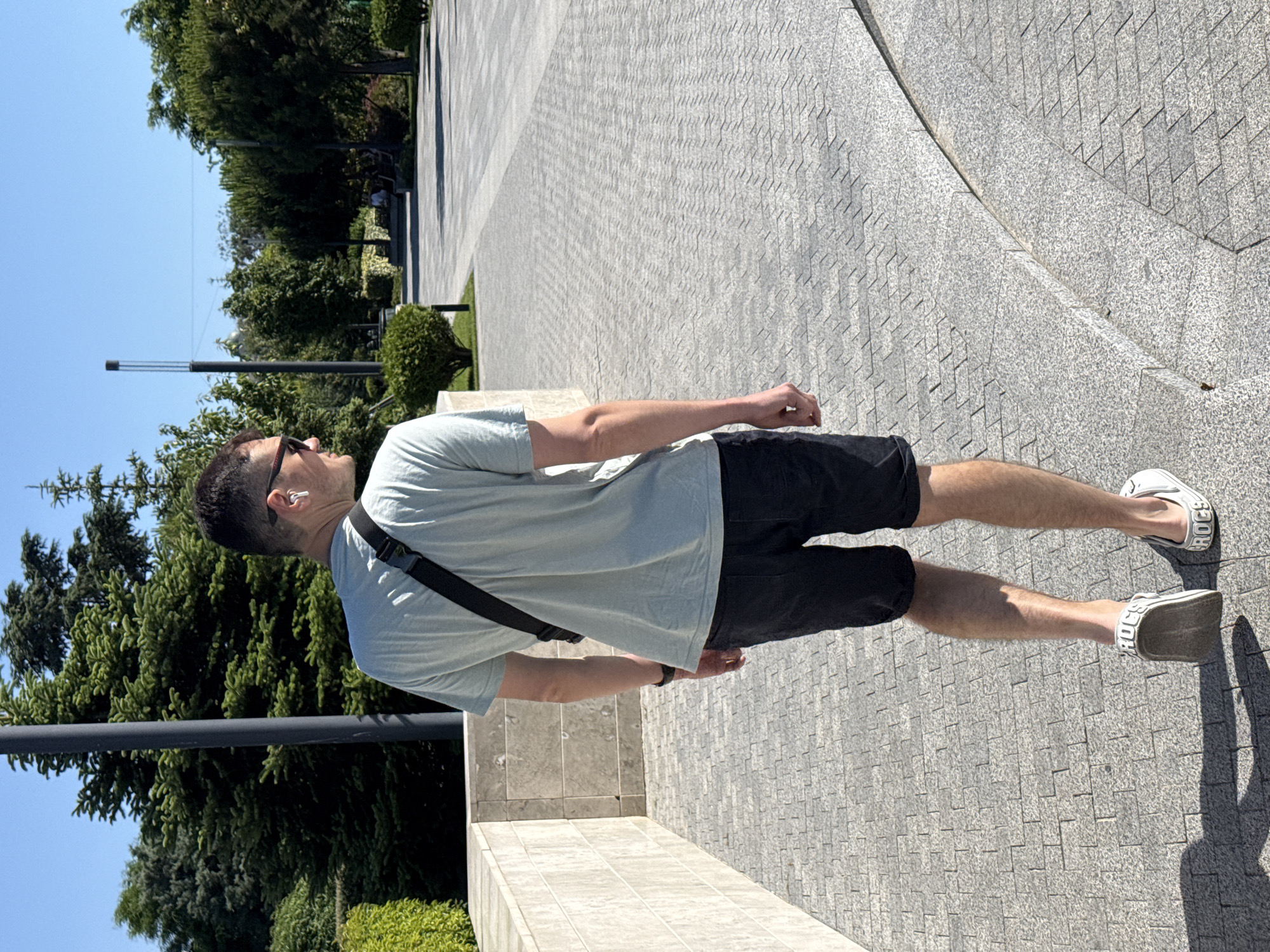A full-body, rear view, medium shot shows a man with short dark hair, wearing sunglasses, a light blue t-shirt, black shorts, and white shoes with the word "CROCS" in black lettering. He is walking away from the camera on a paved pathway in a park on a sunny day. He has a black crossbody bag slung over his right shoulder and is wearing white earbuds. The background consists of trees, bushes, and a stone wall. The paving stones are a light gray color with a geometric pattern. There is a shadow of the man on the ground to his left.