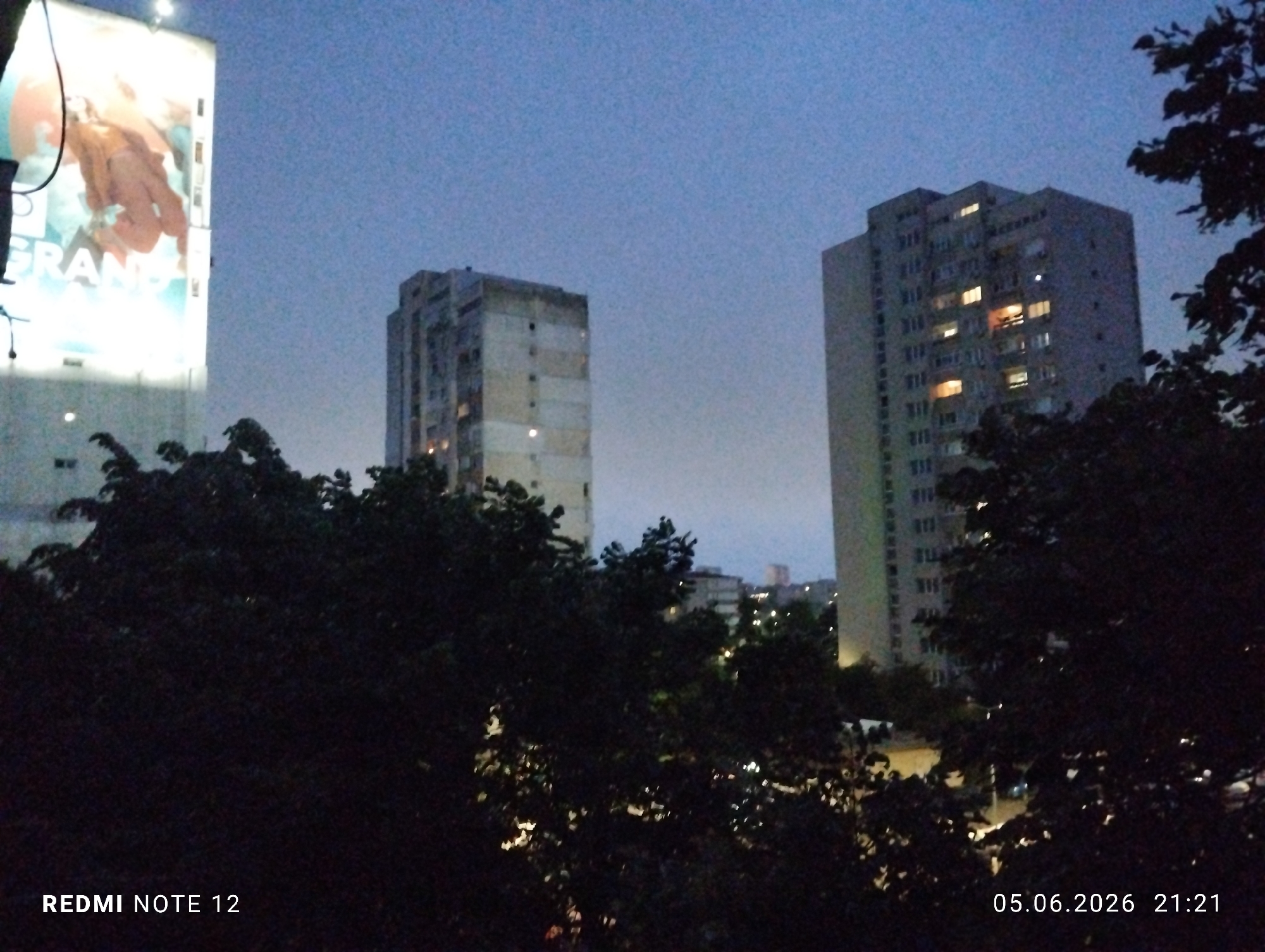 The image displays an urban nocturnal scene in the city of Burgas, Bulgaria, captured at 21:21 on June 5, 2026, using a Redmi Note 12 device. The foreground is dominated by dense, dark foliage, primarily trees and bushes, which obscure the lower sections of the scene and cast deep shadows. Through and above this foliage, several artificial light sources are visible as blurred points of light, suggesting streetlights or distant building illuminations.
In the mid-ground, two prominent multi-story residential buildings are visible against a twilight sky. To the left, a rectangular, light-colored concrete building appears older, with a somewhat weathered facade. A few windows on its visible floors are illuminated with warm interior lights. To the right, a taller, more slender concrete residential building stands, featuring numerous windows, many of which are illuminated with warm light, indicating active habitation. Some windows reveal balconies.
On the far left, a large, brightly illuminated vertical billboard is prominent. It displays a partial advertisement, including the text "GRAN" and a stylized image of a person, possibly a woman, rendered in warm, reddish-orange tones against a lighter background. The billboard's illumination provides a strong light source in the upper-left quadrant of the image.
The sky, visible above the buildings, is a deep, muted blue, characteristic of dusk or early night, and shows a slight gradient from darker blue at the top to a lighter, hazy blue-grey near the horizon. No moon or stars are discernible. There are no clearly visible individuals or discernible actions beyond the implied presence within the illuminated residential units. The overall environment is quiet and residential.