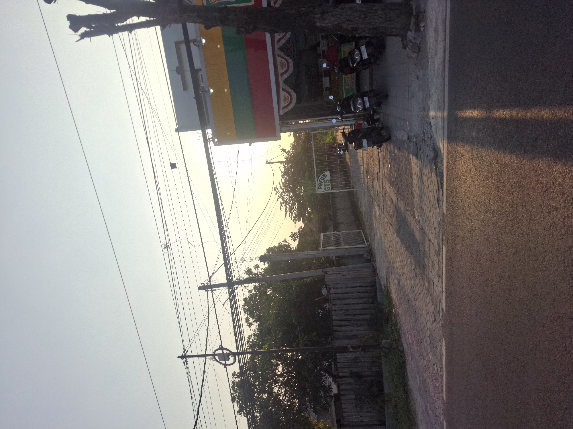 In Banjarmadu, Indonesia, the image depicts a street scene at sunset. A paved road with a white line runs across the foreground, leading towards a sidewalk made of interlocking pavers. To the left of the sidewalk, a wooden fence encloses a property with dense foliage behind it, punctuated by utility poles and numerous overhead wires. A metal gate, labeled "Perum GTS," stands open, leading into the property. To the right of the gate is a building with a brightly colored awning, featuring horizontal stripes of yellow, green, and red. Several motorcycles are parked on the sidewalk in front of the building. The sky is a pale orange, characteristic of a sunset, and a silhouetted tree stands on the right edge of the frame.