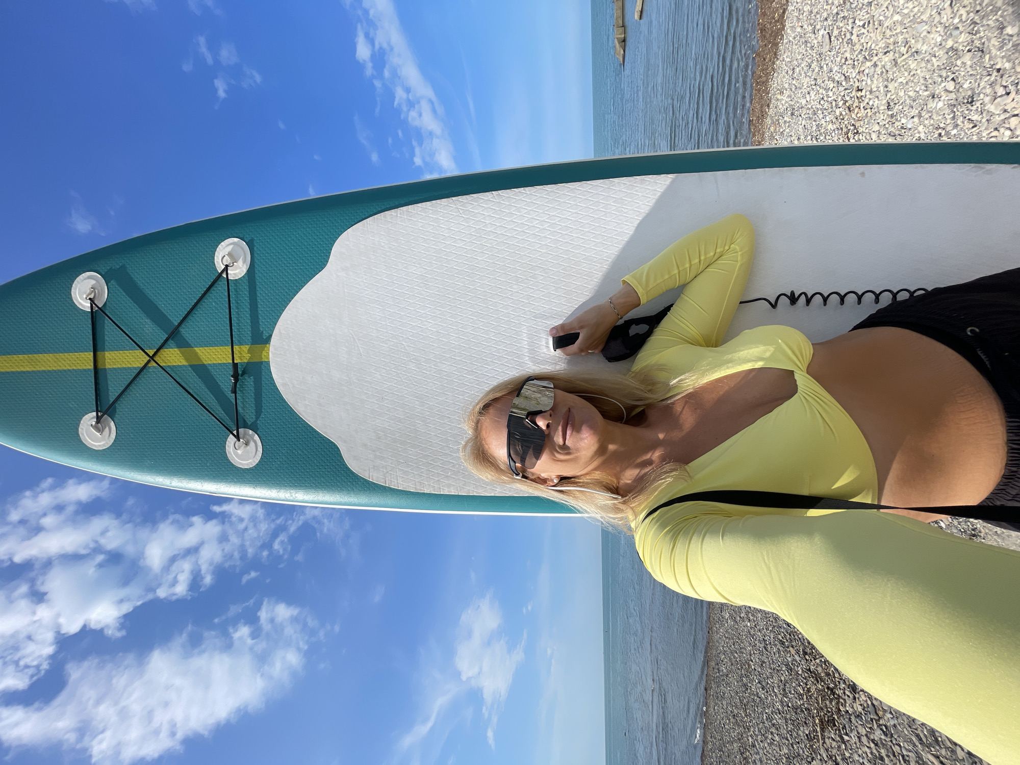 A woman stands on a pebble beach next to a paddleboard. She is wearing a yellow long-sleeved crop top, black shorts, and large, wraparound sunglasses. The paddleboard is green and white with yellow accents and black bungee cords. The ocean is visible in the background, with a clear blue sky and scattered clouds above. The image appears to be a selfie taken by the woman, who is holding the phone out to capture herself and the paddleboard. The location is the city of Sochi, Russia.