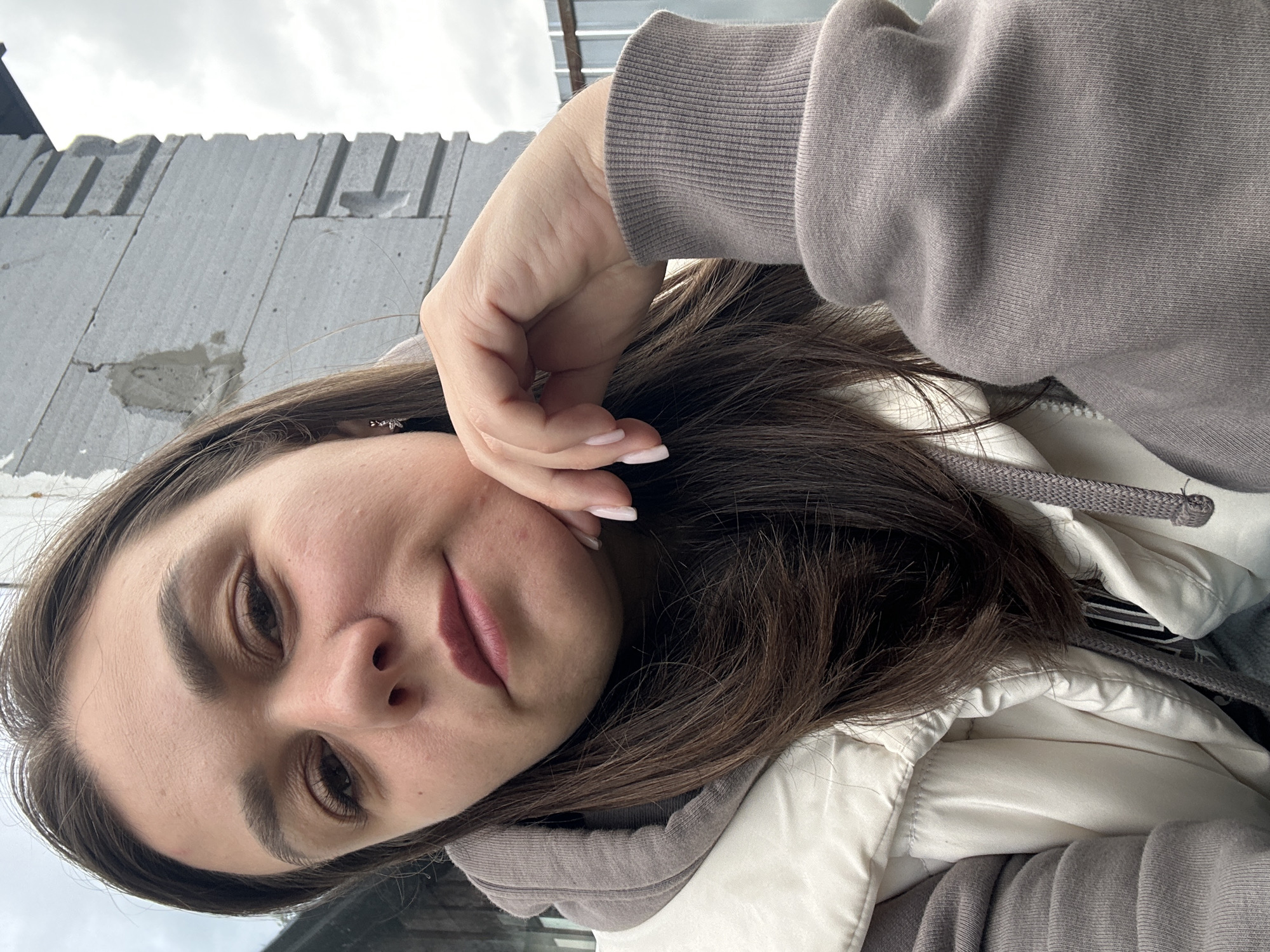 The image is a selfie taken outdoors, likely in a residential or construction area in Chaikovsky, Russia. A woman with long, dark brown hair is the central subject. She is wearing a grey hooded sweatshirt and a cream-colored puffer vest. Her left hand is resting on her chin, and she is looking towards the camera with a neutral expression. In the background, a light grey, unfinished brick wall is visible, possibly part of a building under construction. The sky is overcast.