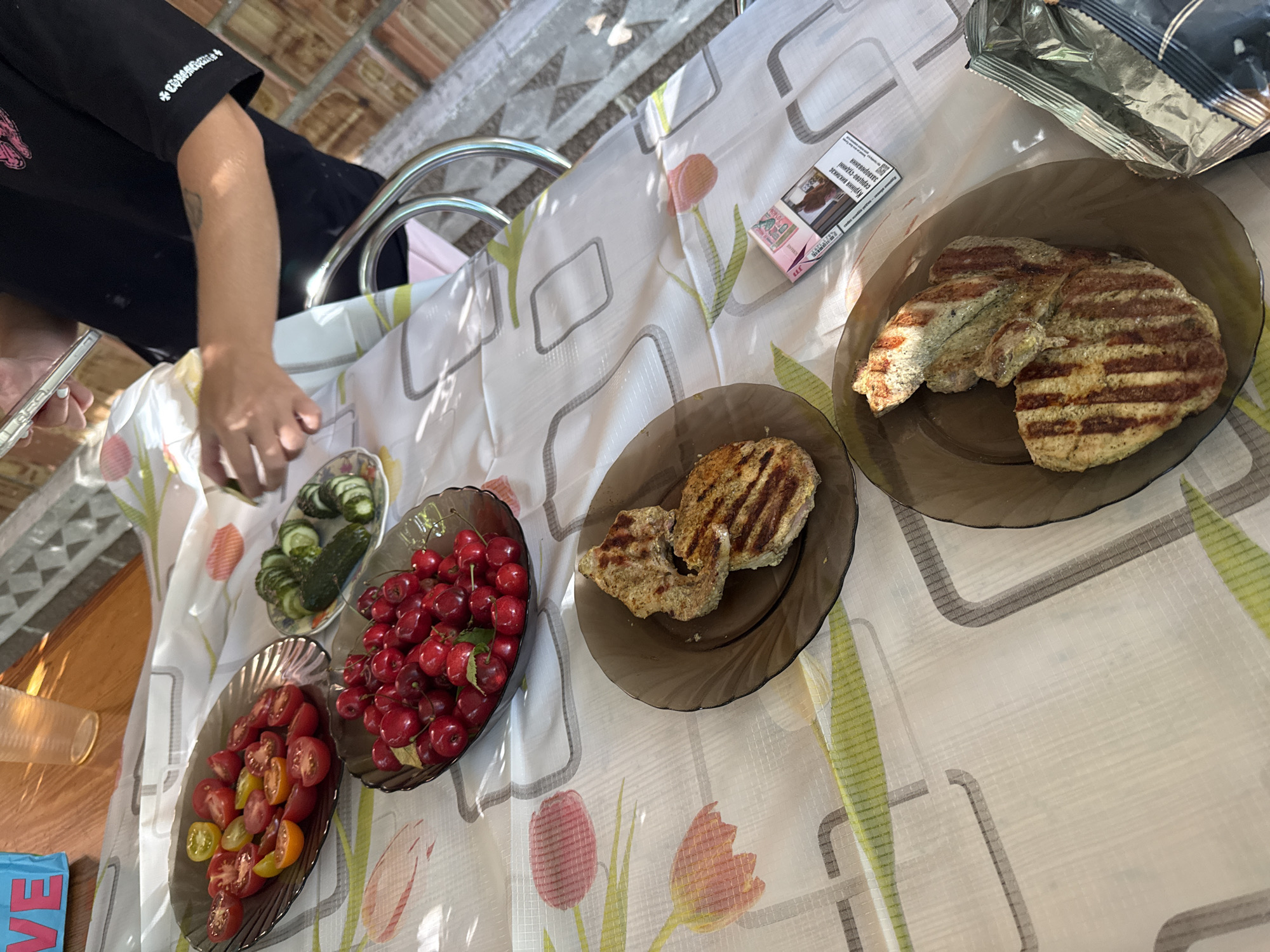 A still life shot taken outdoors on a table covered with a floral-patterned tablecloth depicts a meal. In the foreground, two plates of grilled meat patties are visible. The top plate holds two patties, one slightly overlapping the other, both exhibiting prominent grill marks. The bottom plate contains a single larger patty, also well-grilled. To the right of these plates, a pack of cigarettes and a foil bag are placed on the tablecloth. In the background, a shallow dish of red cherries and another dish containing sliced cherry tomatoes and a whole cucumber with sliced rounds are arranged. A clear plastic cup and a blue bag with "LOVE" written on it are also present. A person's arm and shoulder, wearing a black t-shirt with a graphic design and sleeve embellishments, are visible in the upper right portion of the frame, with their hand holding a smartphone and seemingly interacting with food. The lighting suggests an outdoor setting, possibly in a yard or patio area. The scene is likely in the city of Obukhivka, Ukraine, given the user’s location context.