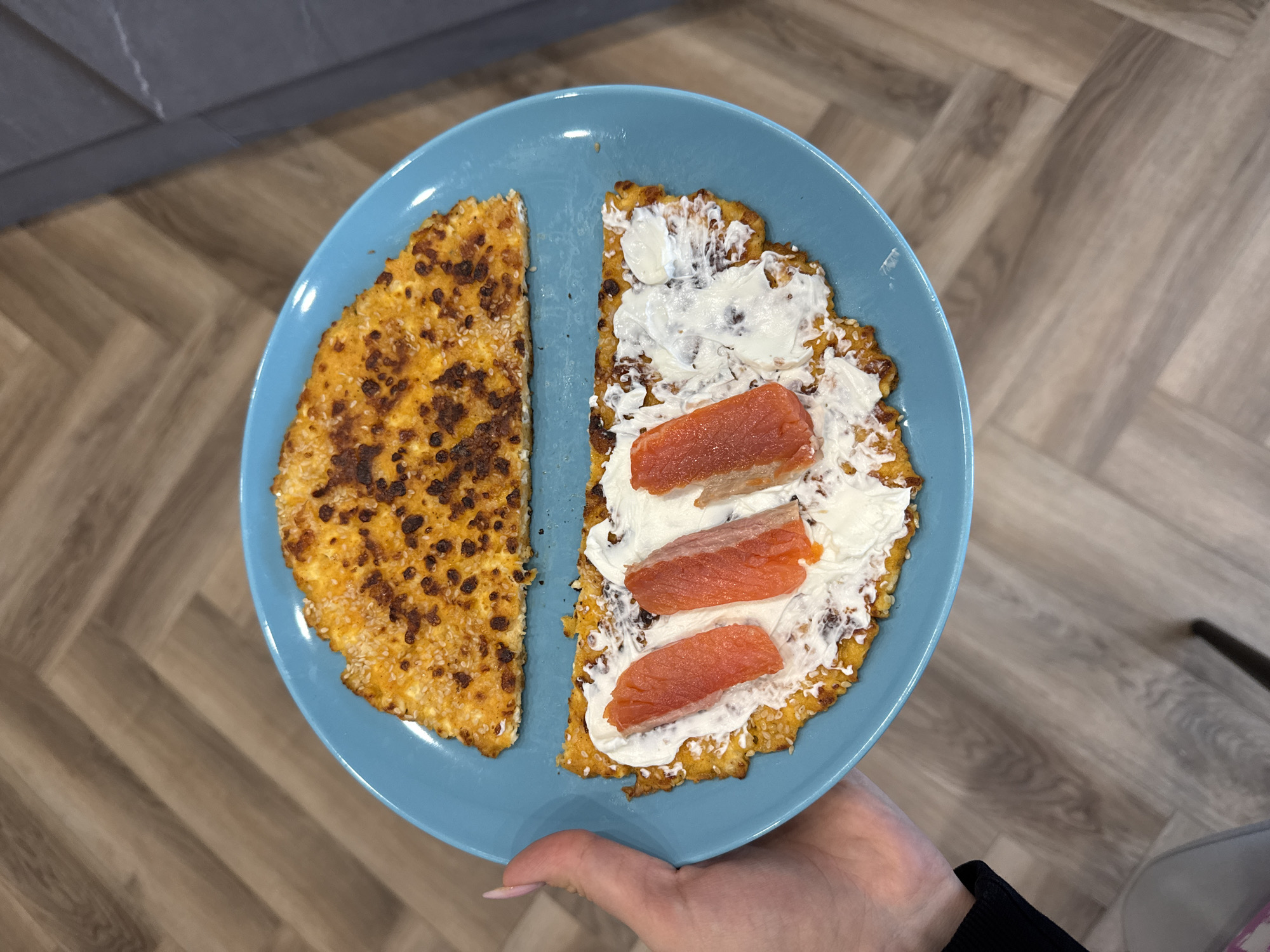 A high-angle, close-up shot shows a person holding a light blue, circular plate filled with food. The plate contains two halves of a large, flat, cooked item, likely a savory pancake, fritter, or flatbread. The left half is plain, displaying a golden-brown, somewhat mottled surface with darker, browned spots and scattered white sesame seeds.
The right half of the food is spread generously with a white, creamy substance, resembling cream cheese or sour cream. Placed on top of the creamy spread are three distinct, elongated pieces of pinkish-orange fish, most likely smoked salmon. The texture of the cooked base under the toppings appears similar to the plain half, also showing some sesame seeds along its edges.
The plate is held by a hand with light-colored nail polish on the fingernails. In the background, the setting appears to be indoors, likely a kitchen or dining area. The floor beneath the plate features light-colored wooden planks arranged in a herringbone pattern. In the upper left corner, a dark grey or charcoal-colored surface, possibly a countertop or cabinet, is partially visible. A dark, thin object, possibly a chair leg, is partially visible in the bottom right corner. There is no visible text in the image.