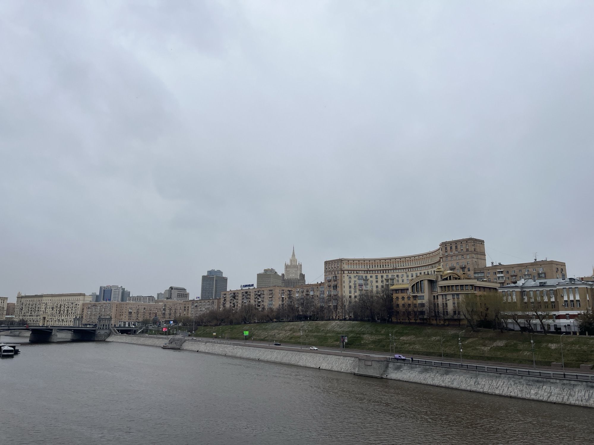 The image shows a cityscape in Moscow, Russia, taken on an overcast day. The Moscow River flows horizontally across the scene, bordered by a concrete embankment. On the far side of the river, there is a roadway with a few vehicles visible. Buildings line the riverbank, ranging in height and architectural style. One prominent structure is a Stalinist skyscraper, likely the Ministry of Foreign Affairs building. Other buildings include residential blocks and commercial structures. A bridge crosses the river on the left side of the image. No people are clearly visible. The overall atmosphere is muted due to the gray sky.