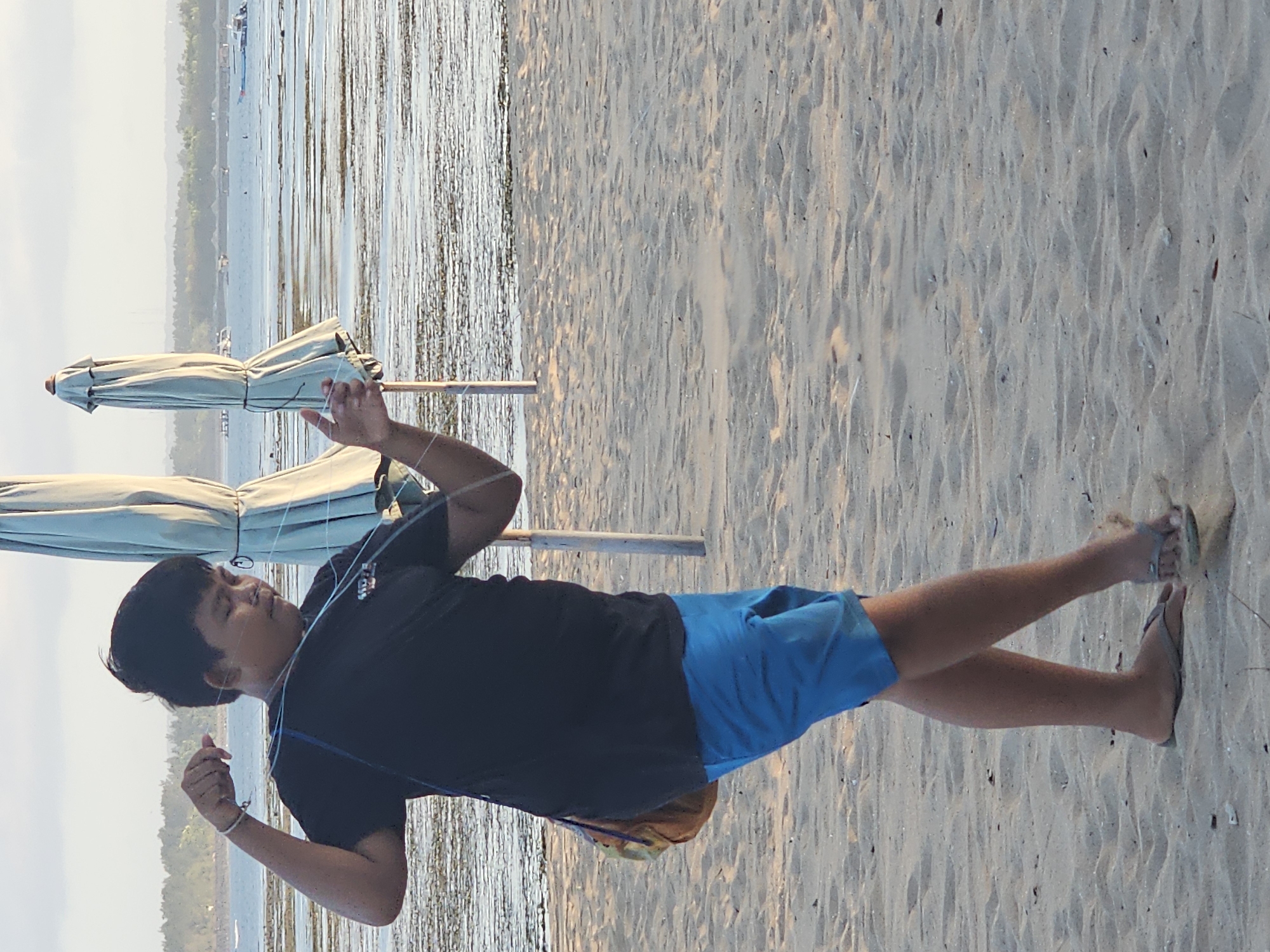 A person is walking on a sandy beach near the ocean. The person is wearing a black t-shirt and blue shorts, with a bag slung over their shoulder. They are holding a string that is attached to something out of frame. In the background, there are two closed beach umbrellas and the ocean with some boats in the distance. The sky is overcast. This scene is likely located in Denpasar, Indonesia, given the context provided.
