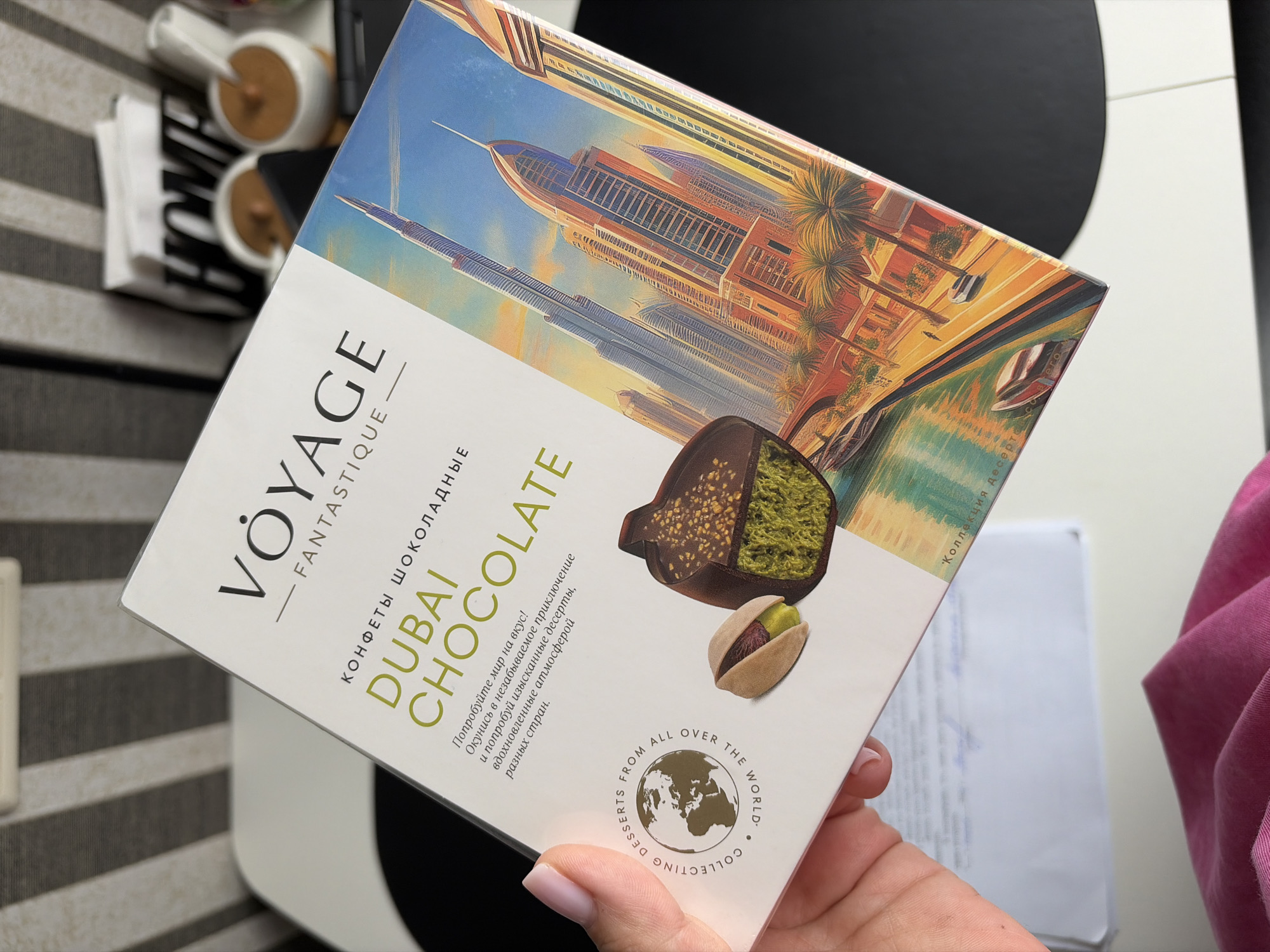 The image shows a box of "VÔYAGE FANTASTIQUE" chocolates with a "DUBAI CHOCOLATE" theme. A hand with light pink nail polish is holding the box. The box features an illustration of Dubai's skyline with skyscrapers and palm trees, a canal with boats, and a close-up of a chocolate with pistachio filling and a whole pistachio. Text on the box reads "КОНФЕТЫ ШОКОЛАДНЫЕ DUBAI CHOCOLATE" and "Попробуйте мир на вкус! Окунитесь в незабываемое приключение и попробуйте изысканные десерты, вдохновленные атмосферой разных стран." and "COLLECTING DESSERTS FROM ALL OVER THE WORLD." There is also a partial view of a white envelope with some blue writing on it. The background is blurred, showing striped patterns and the word "HOME" on decorative objects. The image does not depict the city of Sochi, Russia.