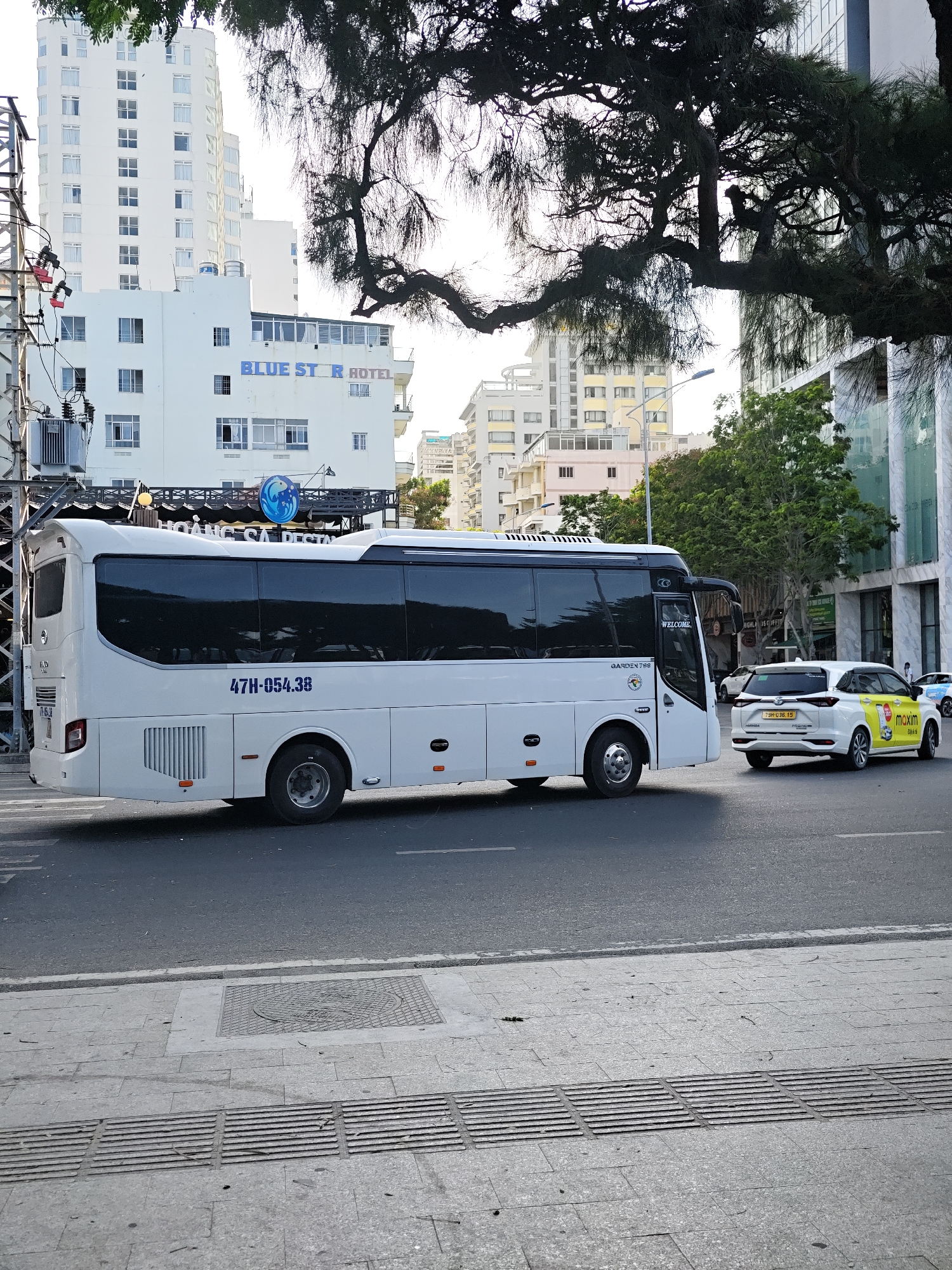 In Nha Trang Ward, Vietnam, a white bus with the license plate "47H-054.38" is captured in motion on a city street, partially obscured by the shadow of a large tree. To the right of the bus, a white car, identified as a "maxim" taxi with the license plate "72H-036.15", is also traveling on the road. In the background, a multi-story white building prominently displays the signage "BLUE STAR HOTEL". The street scene includes a sidewalk with tactile paving, a manhole cover, and various electrical infrastructure on the left side of the image. The sky is bright, suggesting daytime.