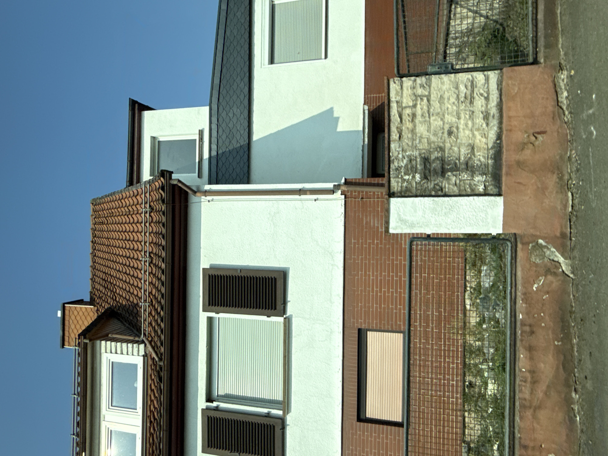 The image shows the exterior of a multi-story building with a distinctive tiled roof and white stucco walls, located in the city of Bad Sachsa, Germany. The building has multiple windows, some with dark brown shutters and roller blinds, and others with light-colored roller blinds. The lower portion of the building is clad in red brick. A metal fence with a wire mesh and red brick pillars separates the property from the street. A portion of the fence is visible, with a stone pillar to its left. Greenery is visible behind the fence. The sky is a clear, bright blue.