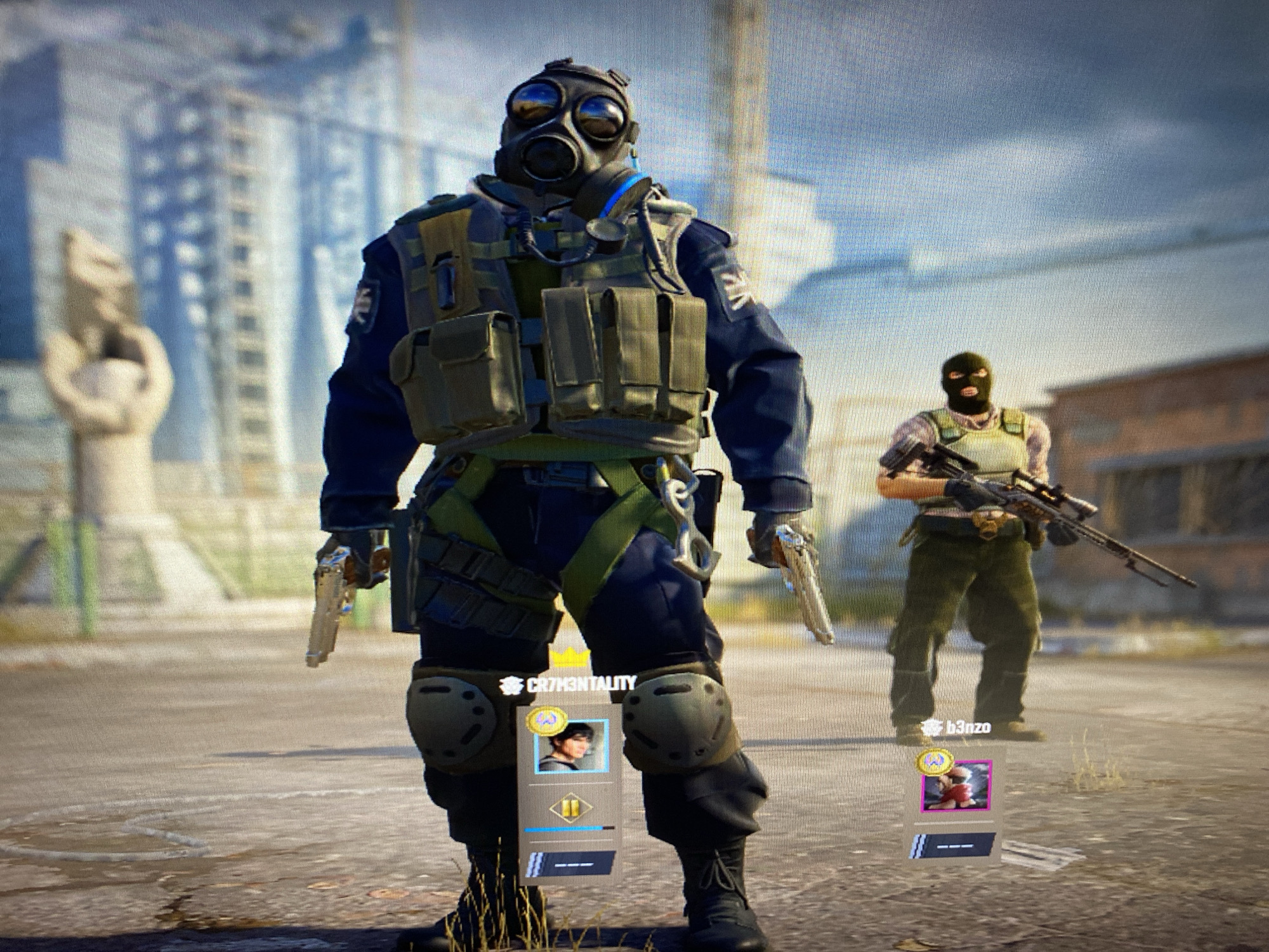 The image is a screenshot from a video game, featuring two male characters in an outdoor, urban, and possibly post-apocalyptic setting during the daytime. The photo exhibits screen pixelation, indicating it was taken of a digital display.
In the foreground, a prominent character stands, clad in a dark blue uniform, a tactical vest with multiple pouches, knee pads, and a full-face gas mask. He holds a silver pistol in each hand, pointed downwards. Below him, a game interface displays the player name "#CR73MENTALITY" with a profile picture of a young man with dark hair.
Further back and to the right, a second, slightly blurred character is visible. This figure wears a black balaclava, a light-colored vest, and green trousers, holding a long rifle, likely a sniper rifle. Beneath this character, the player name "b3nzo" is shown with a profile picture of a female character in red attire.
The environment consists of cracked or worn ground, possibly concrete or asphalt, with sparse dry vegetation. The background features tall, somewhat dilapidated buildings under a clear, light blue sky with scattered clouds. To the left, a large, abstract stone sculpture, resembling a hand holding an object, is partially visible. The characters' gear suggests a combat or survival-themed video game. The provided location context is Yerevan, Armenia.