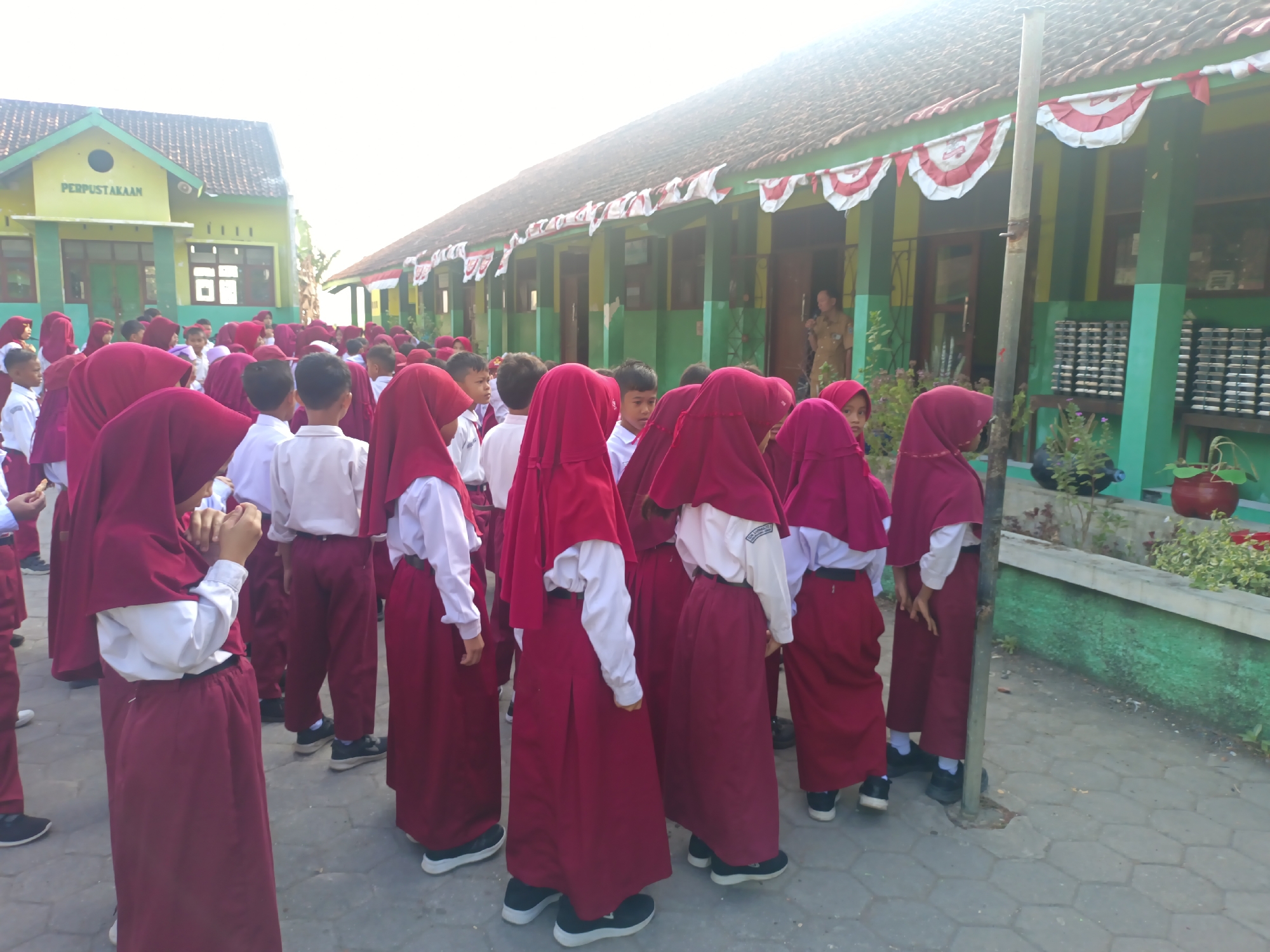 In Sarimulyo, Indonesia, a group of young students wearing maroon hijabs and white shirts stand in a line outside a building. The building, painted yellow and green, has a sign that reads "PERPUSTAKAAN," indicating it is a library. Red and white banners are strung along the building's facade. The ground is paved with grey interlocking stones. One student in the foreground, viewed from the back, wears a maroon hijab and a maroon skirt, with a white shirt underneath. They are looking down at their hands, which are clasped together. Other students in similar uniforms are visible, some facing forward and others with their backs to the camera. The overall scene suggests a school gathering or activity taking place outdoors.