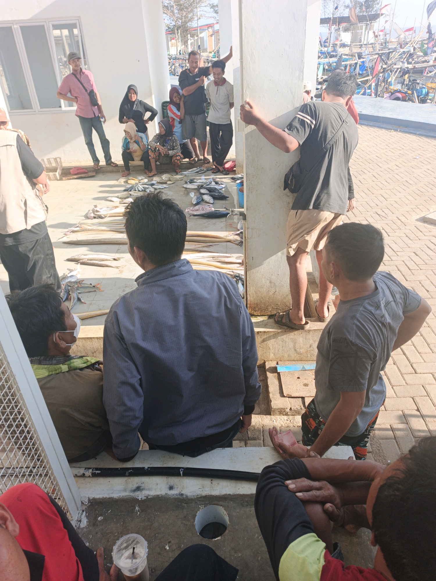 In an outdoor area with a paved ground and a white building in the background, several people are gathered. Some individuals are seated while others stand, and they appear to be engaged in an exchange or transaction. Various types of fish and seafood are spread out on the ground, suggesting a marketplace or a place where fish are being sold or distributed. In the background, there are boats and masts, indicating proximity to a body of water, possibly a harbor or fishing village. The overall scene depicts a lively, open-air environment associated with fishing activities and commerce.