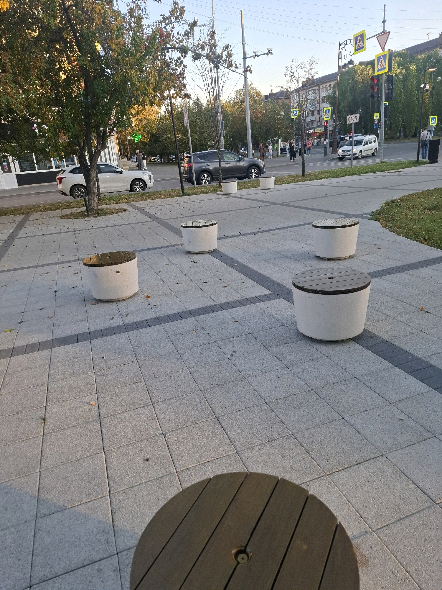 The image depicts a public space in Tyumen, Russia, likely an urban park or pedestrian walkway, as indicated by the presence of concrete benches, tiled paving, and streetlights. Several cylindrical concrete benches with wooden tops are scattered across the paved area. In the background, a street is visible with traffic, including cars and a white van, and a pedestrian crossing with traffic lights and road signs. Buildings line the street, suggesting a city environment. Trees with autumn foliage are present, casting shadows on the ground, implying the season is autumn. A few people are visible in the distance near the street and crosswalk. The overall scene conveys a typical urban setting in Russia during daylight hours.