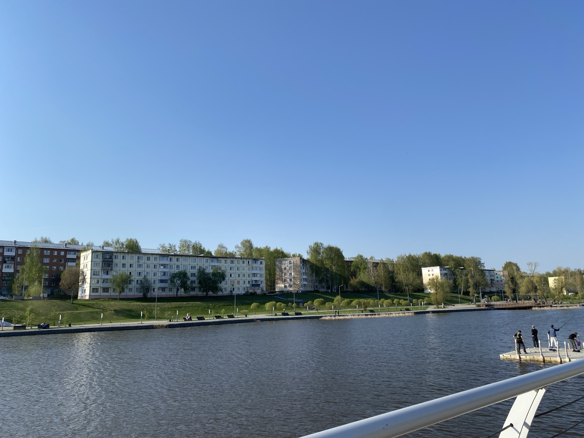 The image captures a view of Votkinsk, Russia. A body of water dominates the foreground, reflecting the clear blue sky. Along the opposite bank, several multi-story apartment buildings line the shore, punctuated by trees with fresh spring foliage. A paved walkway runs along the riverbank, dotted with benches and occasional figures sitting or strolling. In the water, a small floating platform supports a group of individuals engaged in fishing. A metal railing is visible in the lower foreground.
