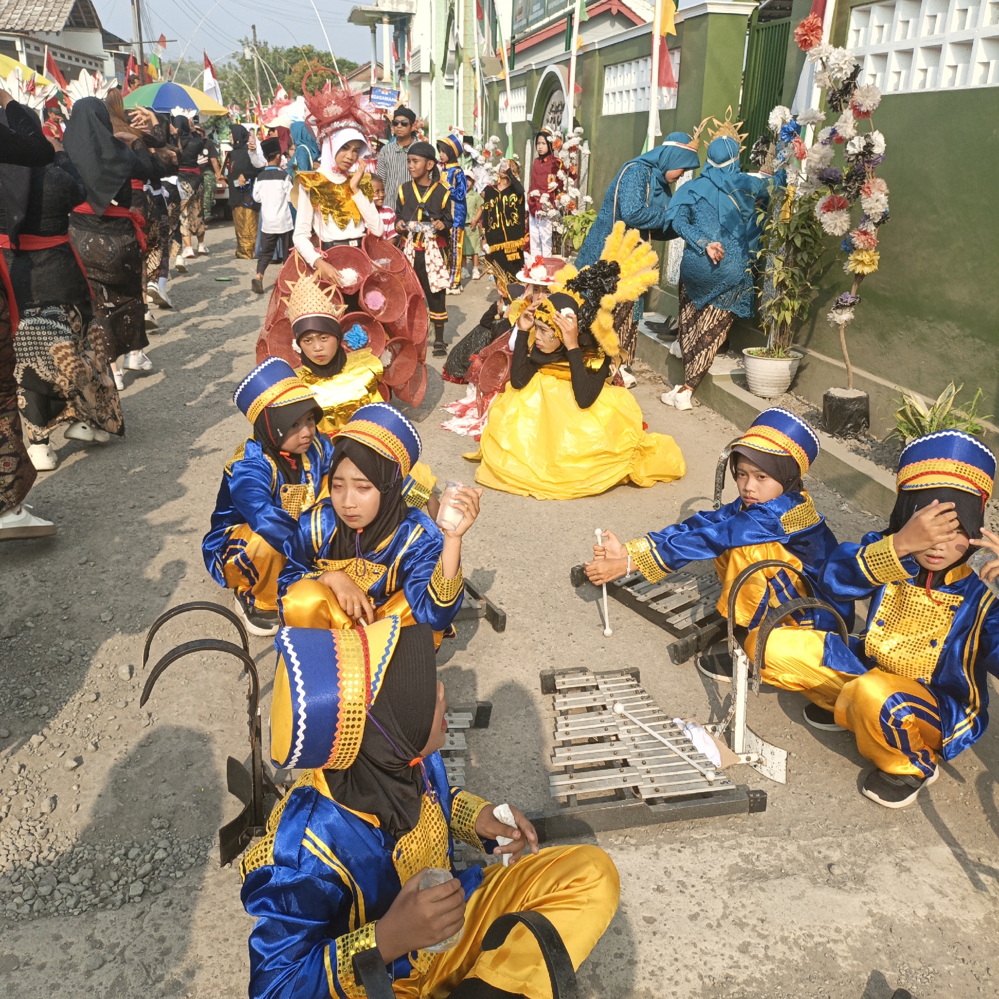 This image depicts a street scene in Tanahbaya, Indonesia, likely during a parade or festival. In the foreground, several children are dressed in matching blue and gold uniforms, some wearing headscarves and elaborate headdresses. Two of the children are seated on the ground, one holding a clear plastic cup and the other resting their hand on their face. A metallophone instrument is positioned on the ground between them, with a drumstick nearby. Other children in similar attire are visible further back in the frame, along with adults and other participants in traditional costumes. The street is paved with gravel, and a building with a green facade and decorative white lattice work lines the right side of the scene. Flags and colorful decorations are visible throughout the background, suggesting a celebratory event.