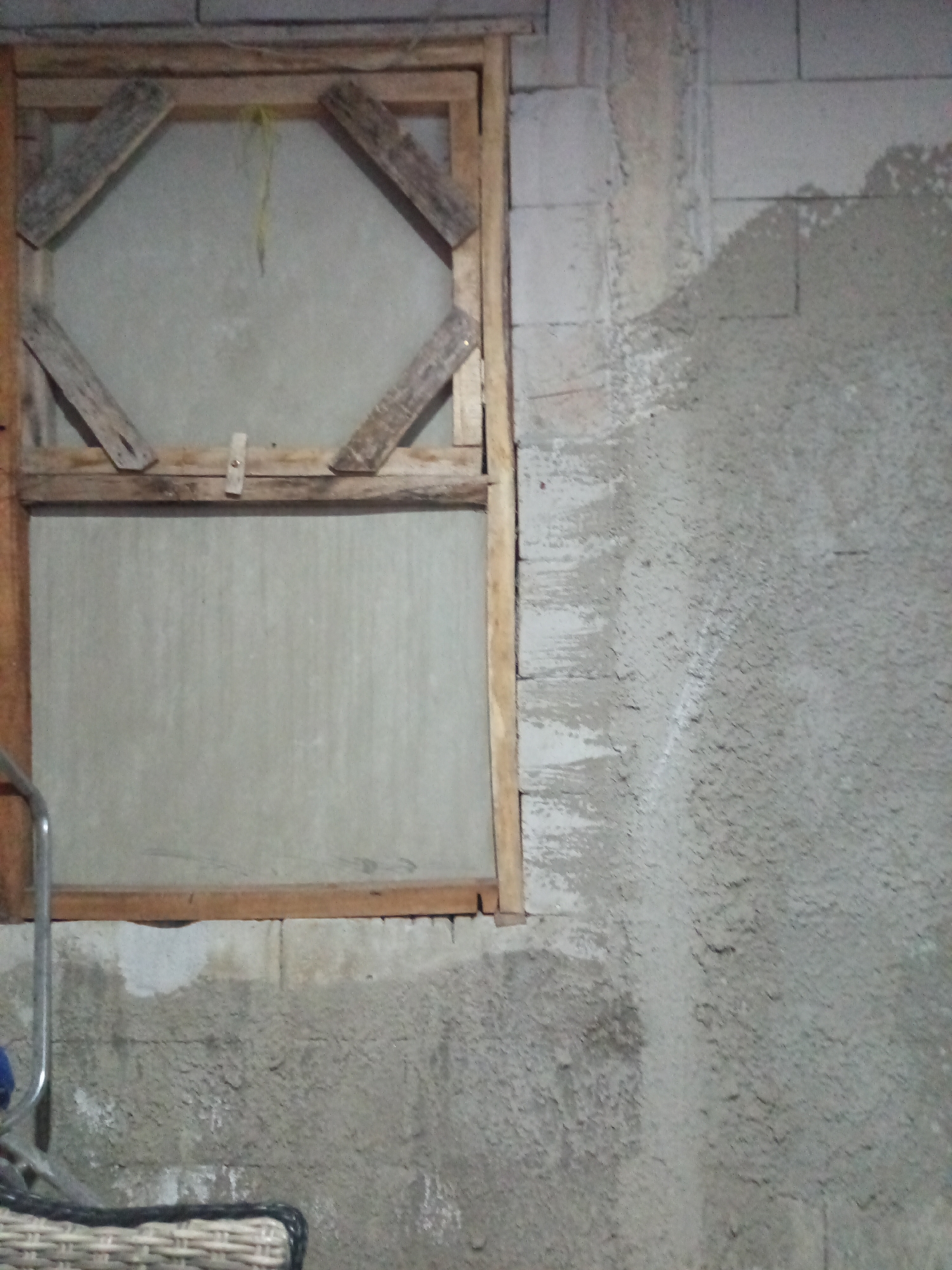 The image depicts a window frame made of rough wood, set against a concrete block wall. The window itself appears to be covered with a light-colored, semi-transparent material. The wooden frame has diagonal pieces of wood attached to the top portion, creating a diamond-like pattern. The wall on the right side of the frame is rough and uneven, showing signs of concrete application. A portion of a metal railing is visible on the left side of the frame, and the edge of a woven basket is in the lower left corner. The overall scene suggests an outdoor or unfinished indoor setting, possibly in a residential or construction area. The lighting appears to be dim.