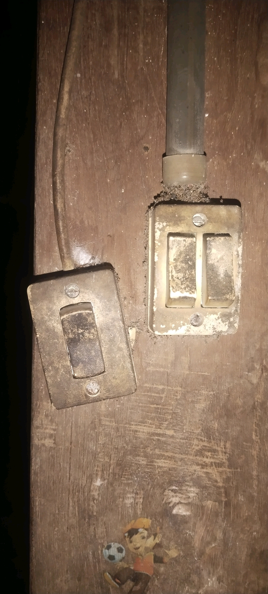 In Daleman, Indonesia, an image captures a close-up view of two old, weathered electrical switches mounted on a rough, wooden surface. The left switch is a single toggle switch with a metallic cover, tilted slightly. The right switch is a double toggle switch, also with a metallic cover, and appears to be covered in a fine layer of dirt or dust. A grey electrical conduit pipe extends downwards from above, connecting to the right switch assembly. A dark, coiled electrical wire is visible to the left, running towards the left switch. The wooden surface shows visible grain and some scuff marks. In the lower portion of the image, a faded decal or sticker depicting a cartoon character playing soccer is adhered to the wood. The overall impression is one of an aged, possibly rural, electrical installation.