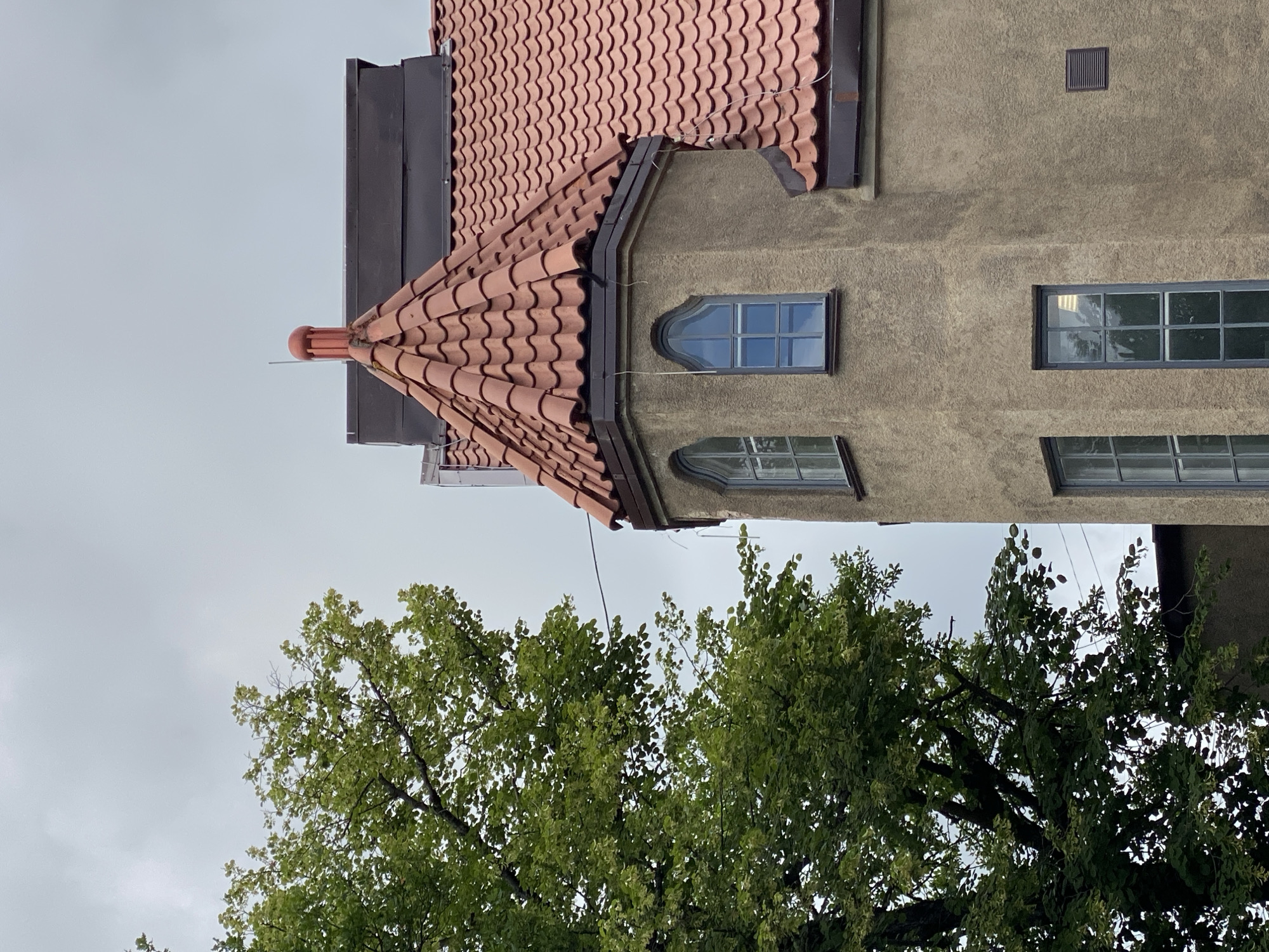The image shows a detail of a tan stucco building with a red tiled roof, featuring a turret with arched windows. A large green tree with dense foliage is positioned to the left of the building, partially obscuring the view of the structure. The sky is overcast with a light gray cloud cover. The scene is likely captured on a day with diffused light, typical of a cloudy or rainy climate. The building's architecture suggests it could be an older structure, possibly in a historical district or a residential area. The presence of the tree and the building's texture create a sense of depth and architectural interest.