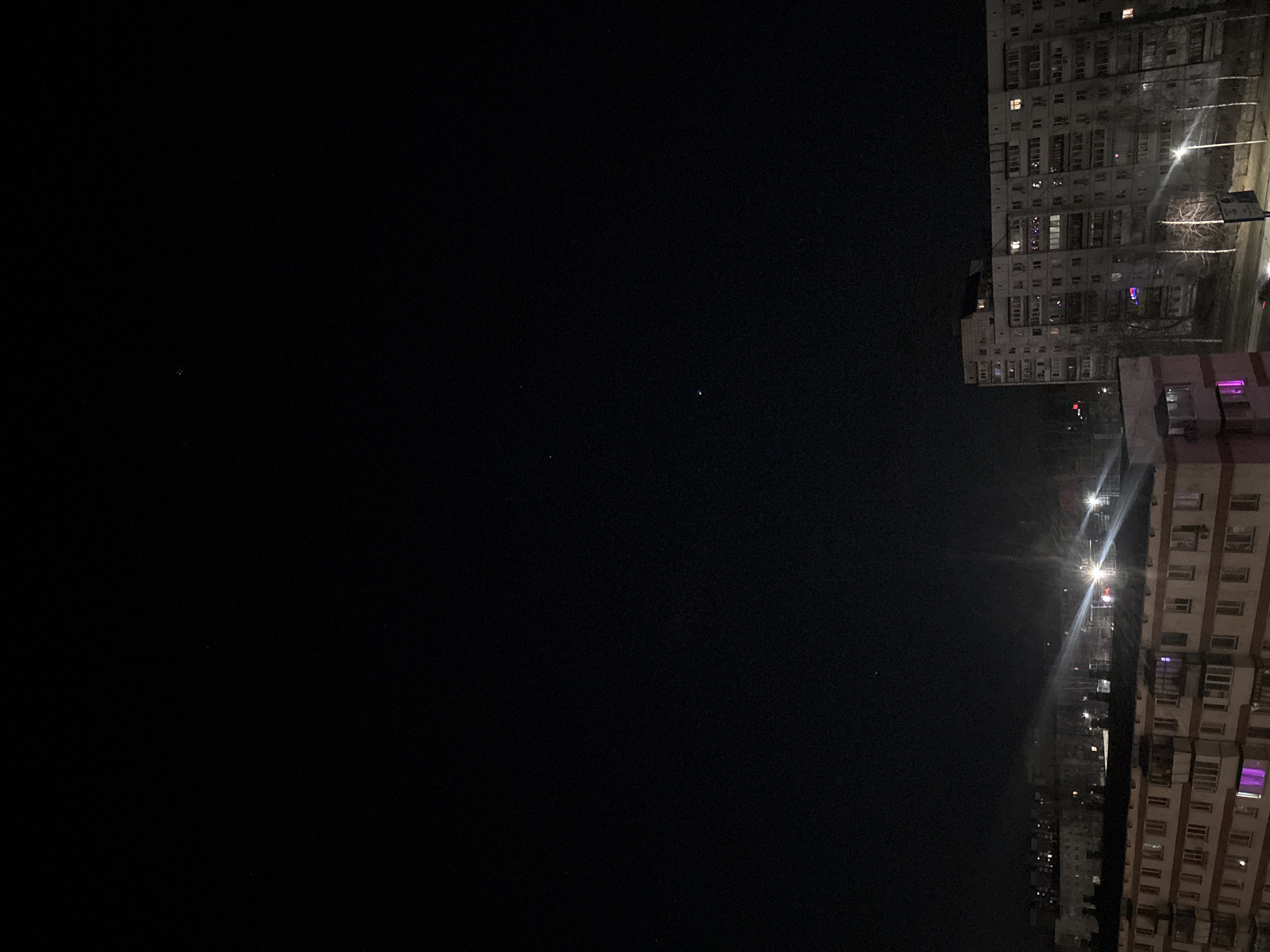 The image depicts a nighttime view from an elevated position in the city of Chaikovsky, Russia. To the right, multiple tall apartment buildings are visible, illuminated by streetlights and lights from within the apartments, some with a distinct purple hue. The left side of the frame is dominated by a dark night sky with several sparsely visible stars. The location can be identified as Chaikovsky, Russia, due to the style of architecture of the apartment buildings being common in this geographical area. No people are visible. No specific actions are taking place in the frame.
