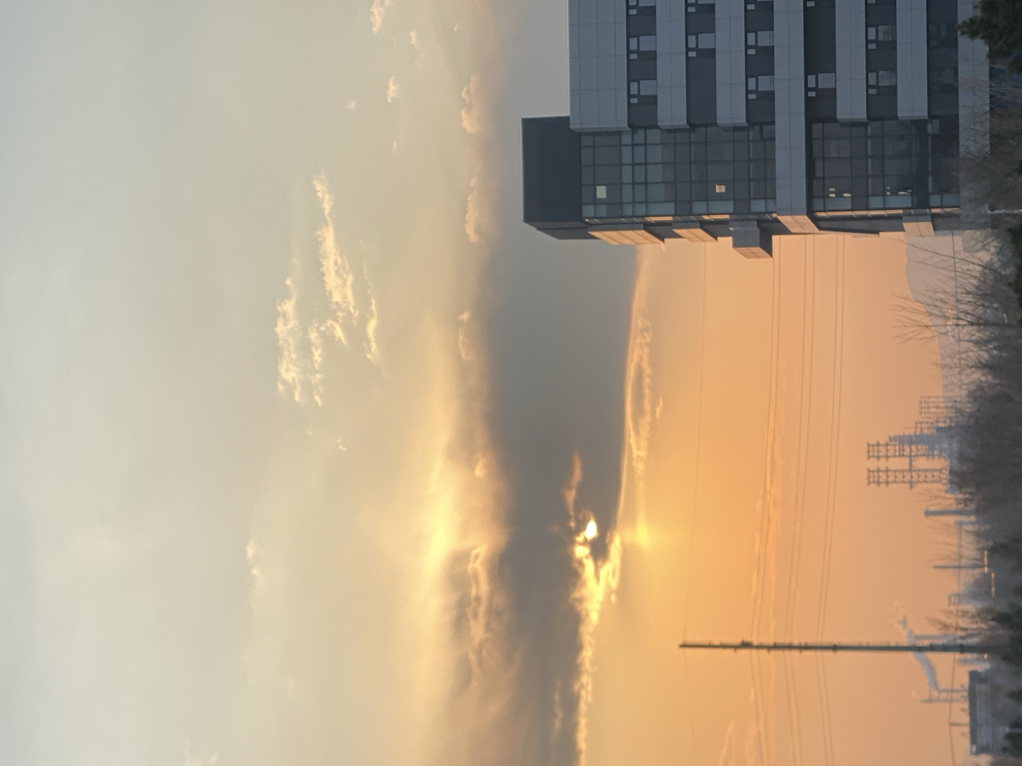 The image depicts a cityscape at sunrise or sunset, likely in the vicinity of Zhongtun, China. The sun is partially obscured by a dark cloud formation with bright rays emanating from behind it. A modern building with a grid-like facade of windows and a grey block stands to the right. Trees and power lines are visible in the background, partially obscured by haze. The sky transitions from light yellow to orange, indicative of sunrise or sunset.