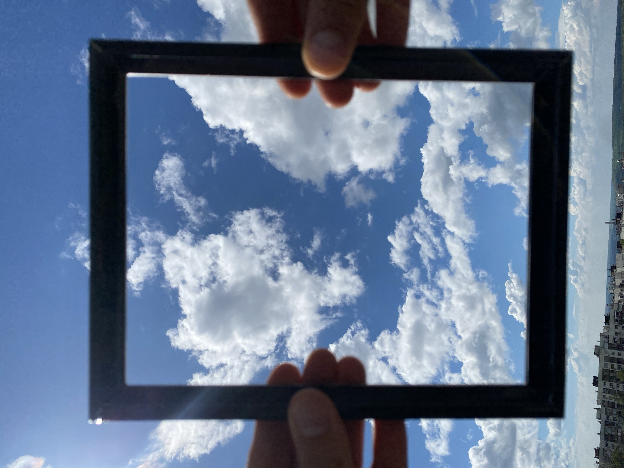A pair of hands holds a rectangular black picture frame horizontally against a bright blue sky filled with white, fluffy cumulus clouds. The hands, with light skin tone, are visible at the top and bottom edges of the frame, firmly grasping it. Through the framed opening, the sky and clouds are prominently featured, displaying varying shades of blue and white, indicating a sunny day with scattered cloud cover.
Outside the frame, particularly on the right side of the image, a distant urban landscape is visible, consistent with the provided location context of Pleven, Bulgaria. This background features a row of multi-story buildings, appearing somewhat hazy and muted against the horizon. A line of greenery, likely trees or a park, separates the distant structures from the vast expanse of the sky. The lighting suggests it is daytime, possibly late morning or early afternoon, with clear weather. The image conveys a creative act of framing a natural scene within an artificial boundary, contrasting the expansive sky with a structured view. There is no visible text in the image.