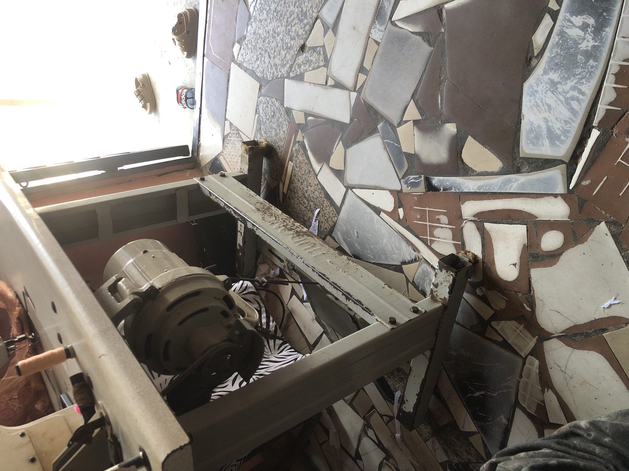 An eye-level shot looks at a sewing machine's motor and table leg. The sewing machine's motor is an olive-green color and is mounted underneath the table. The table itself is made of a light-colored metal with some rust spots and is supported by a metal leg. To the right of the sewing machine, there are three pairs of sandals on the floor, which appears to be tiled with a mosaic pattern. The sandals are of different colors and styles, with one pair appearing to be brown and the other a mix of red, white, and black. The background of the image is blurry and shows a bright, sunlit outdoor area, possibly a doorway or an open space. The image is captured in Ado, Nigeria.
